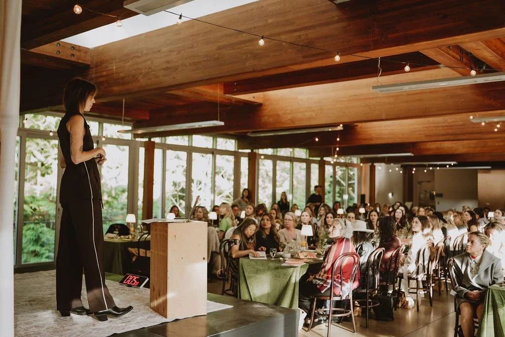 A woman in a black outfit is speaking on a stage in a large room filled with seated audience members. The room has wooden ceiling beams and large windows with greenery outside. The audience is listening attentively, and there are small lamps on the t