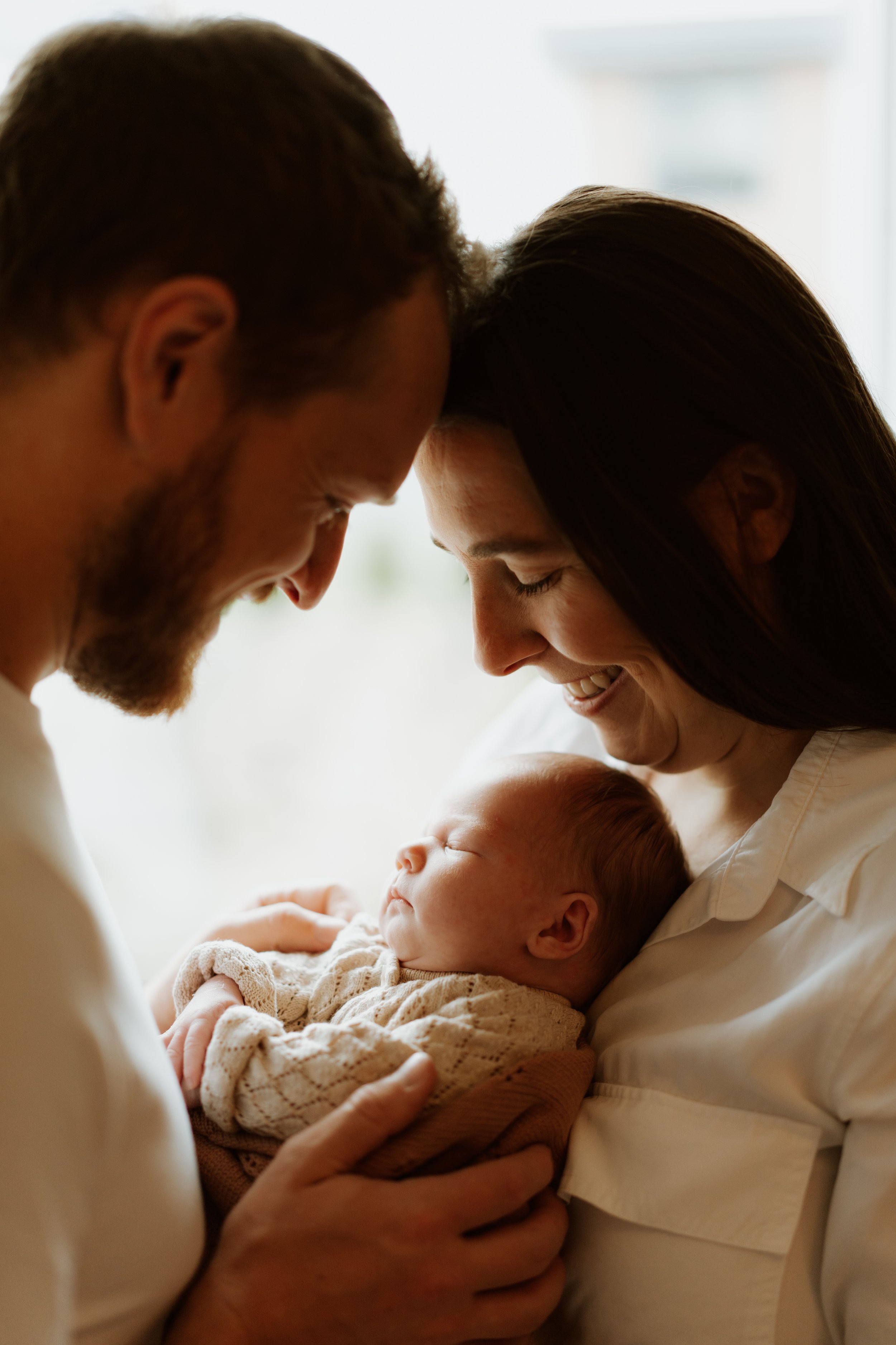 A couple with their heads touching, holding a newborn baby and smiling in a bright room.