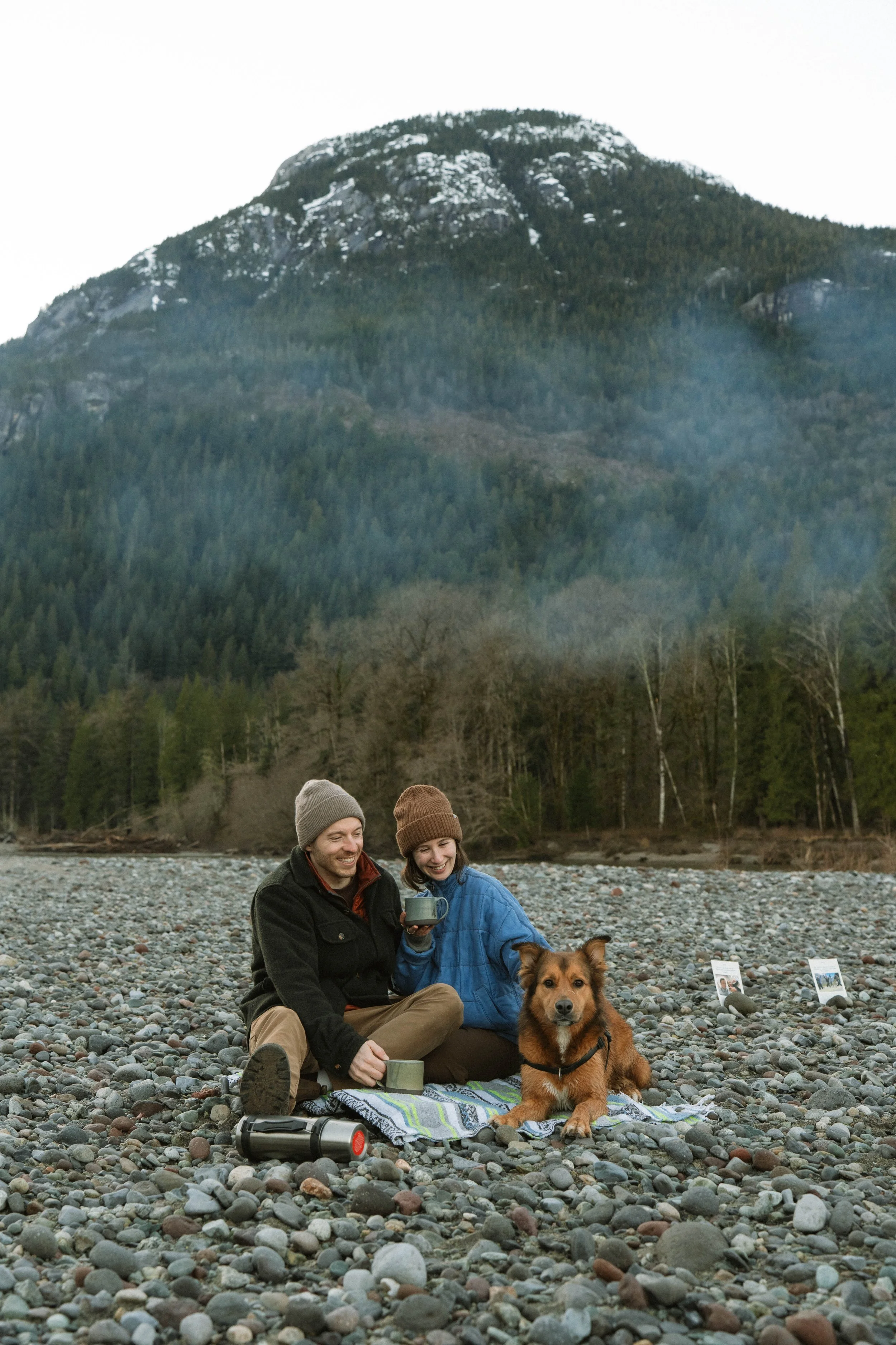 Happy couple sitting on a rocky riverbank with a dog, scenic mountain and forest background, holding mugs, with camping gear nearby.