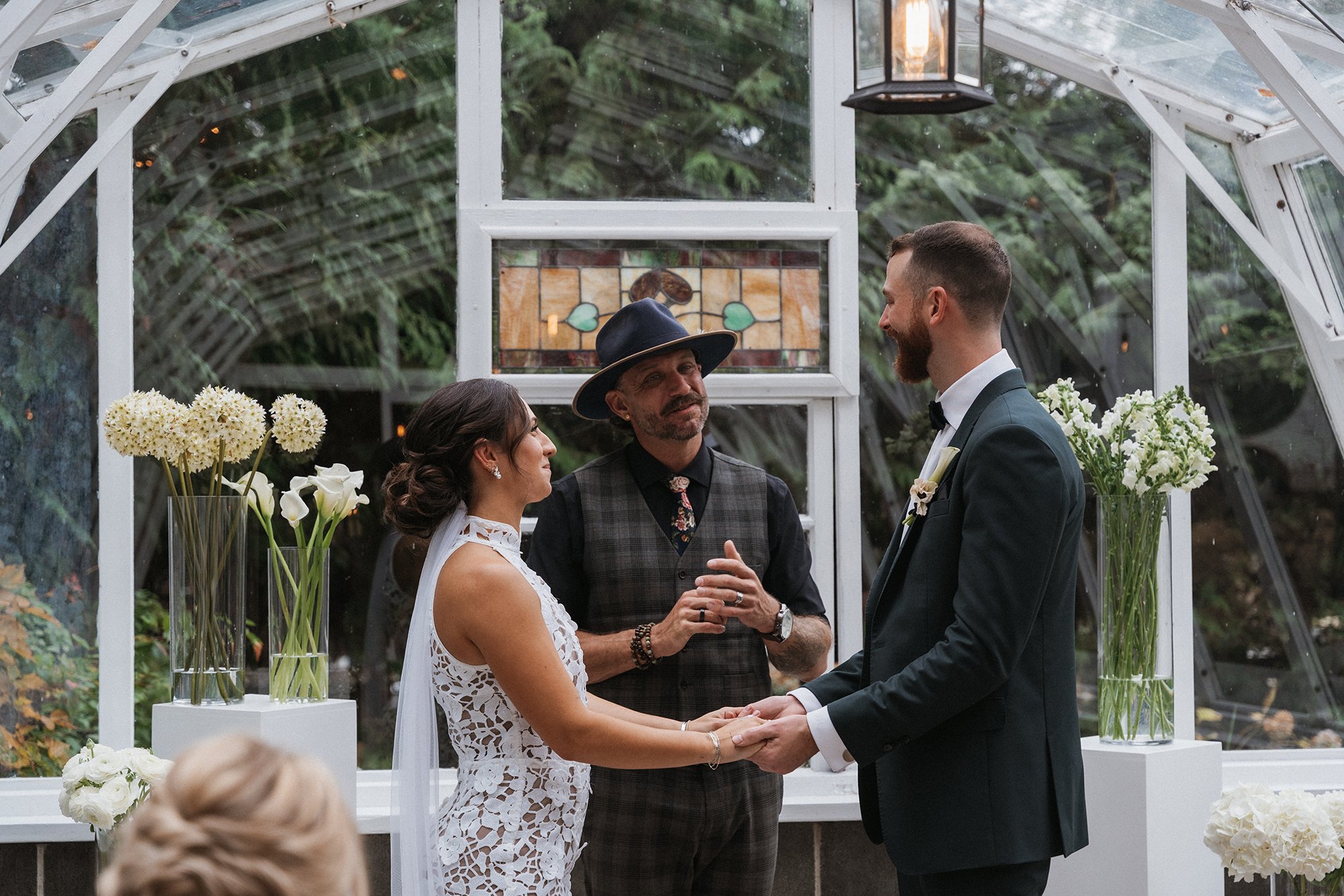 A bride and groom holding hands during their wedding ceremony inside a greenhouse with glass walls and ceiling. A officiant stands between them, holding their hands and speaking. The background shows green plants and a stained glass window. Flower ar