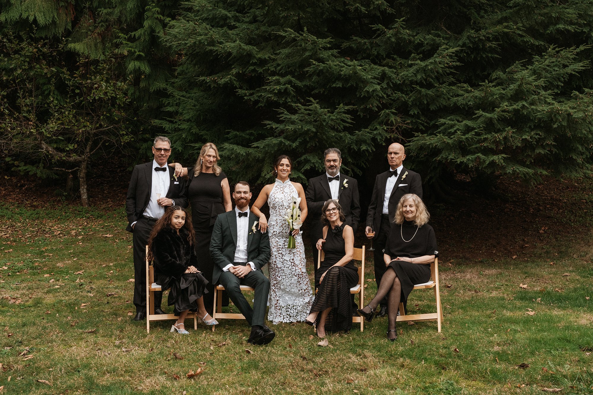 A group of nine people, dressed formally in black-tie attire, gathered outdoors in front of large green trees for a wedding photo. Eight people are standing in the back row, and one woman is seated in front. The bride holds a bouquet, and some people