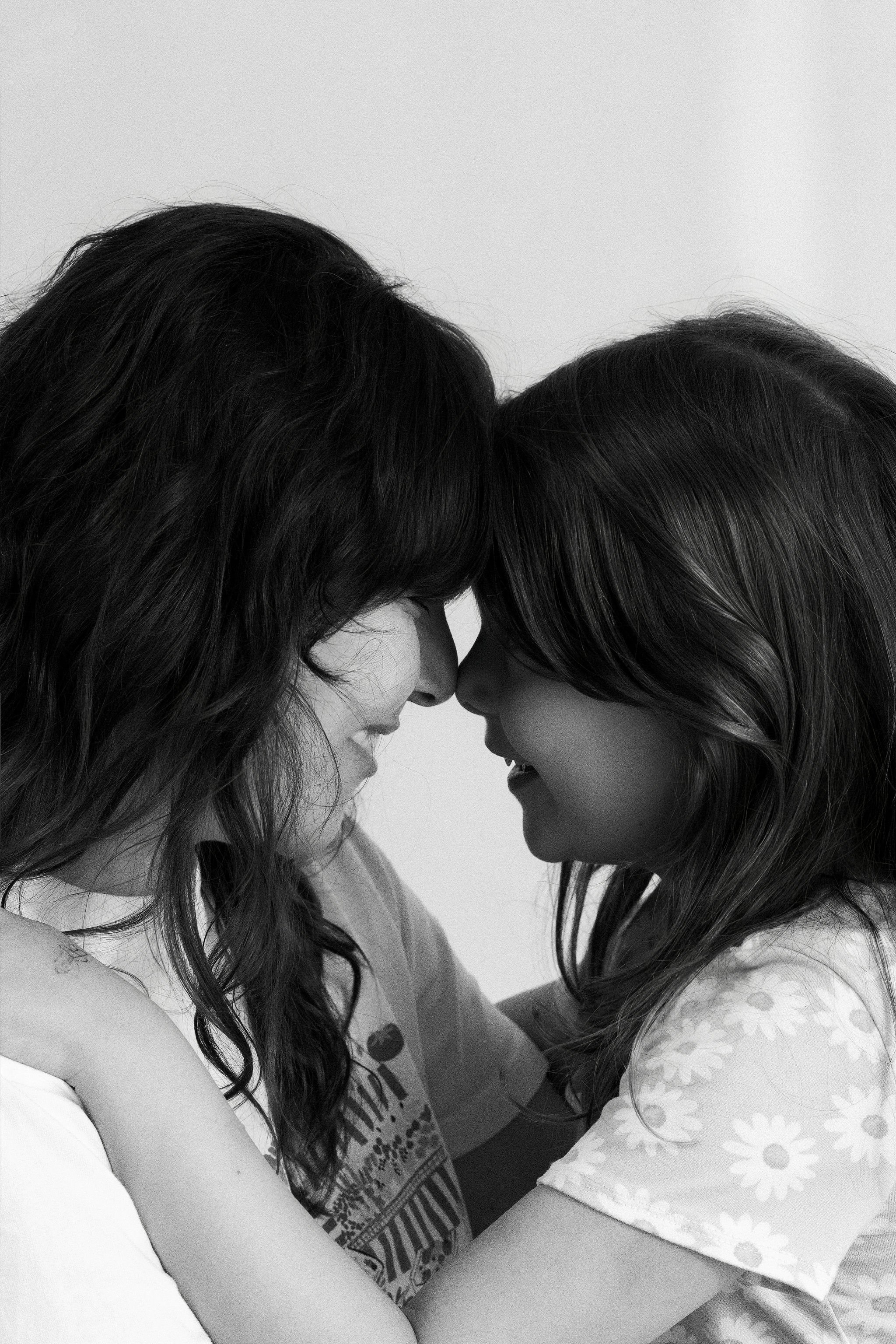 Two women with long hair, one with dark hair and one with light hair, leaning their foreheads together with closed eyes. They are smiling and wearing casual tops, one with a floral pattern and the other with a graphic design. The image is black and w