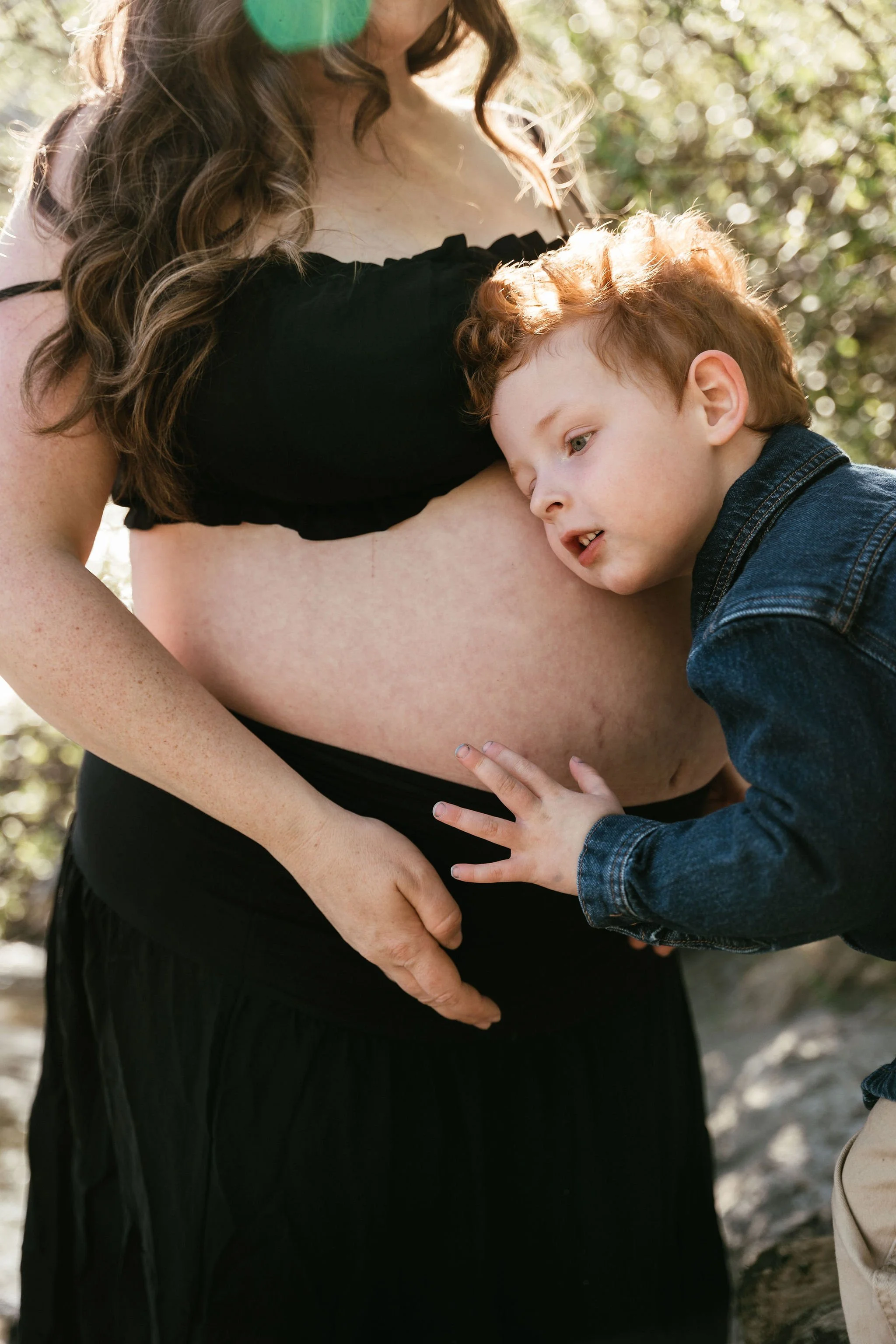A pregnant woman in a black dress with curly hair stands outdoors, with a young boy with red hair and freckles leaning against her belly, touching it, in a sunlit natural setting.