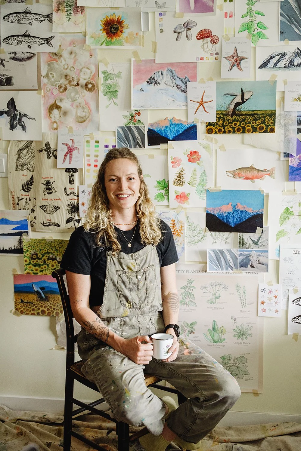 A woman with curly hair sitting on a wooden chair in front of a wall covered with various watercolor paintings and sketches of nature, animals, and landscapes. She is smiling, holding a coffee mug, and wearing paint-splattered overalls over a black t