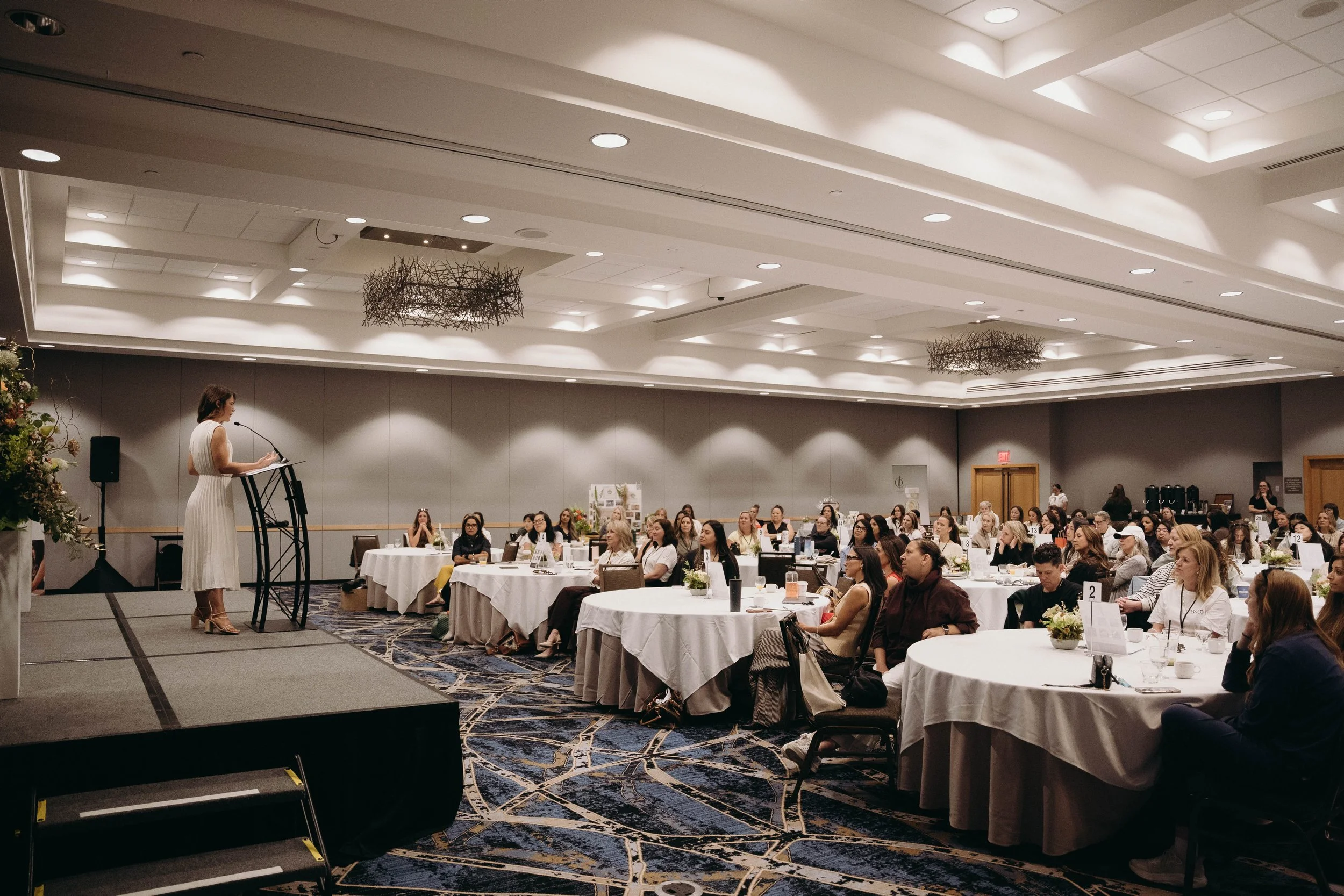 A woman is speaking at a podium to a large audience seated at round tables in a conference room with high ceilings and decorative light fixtures.