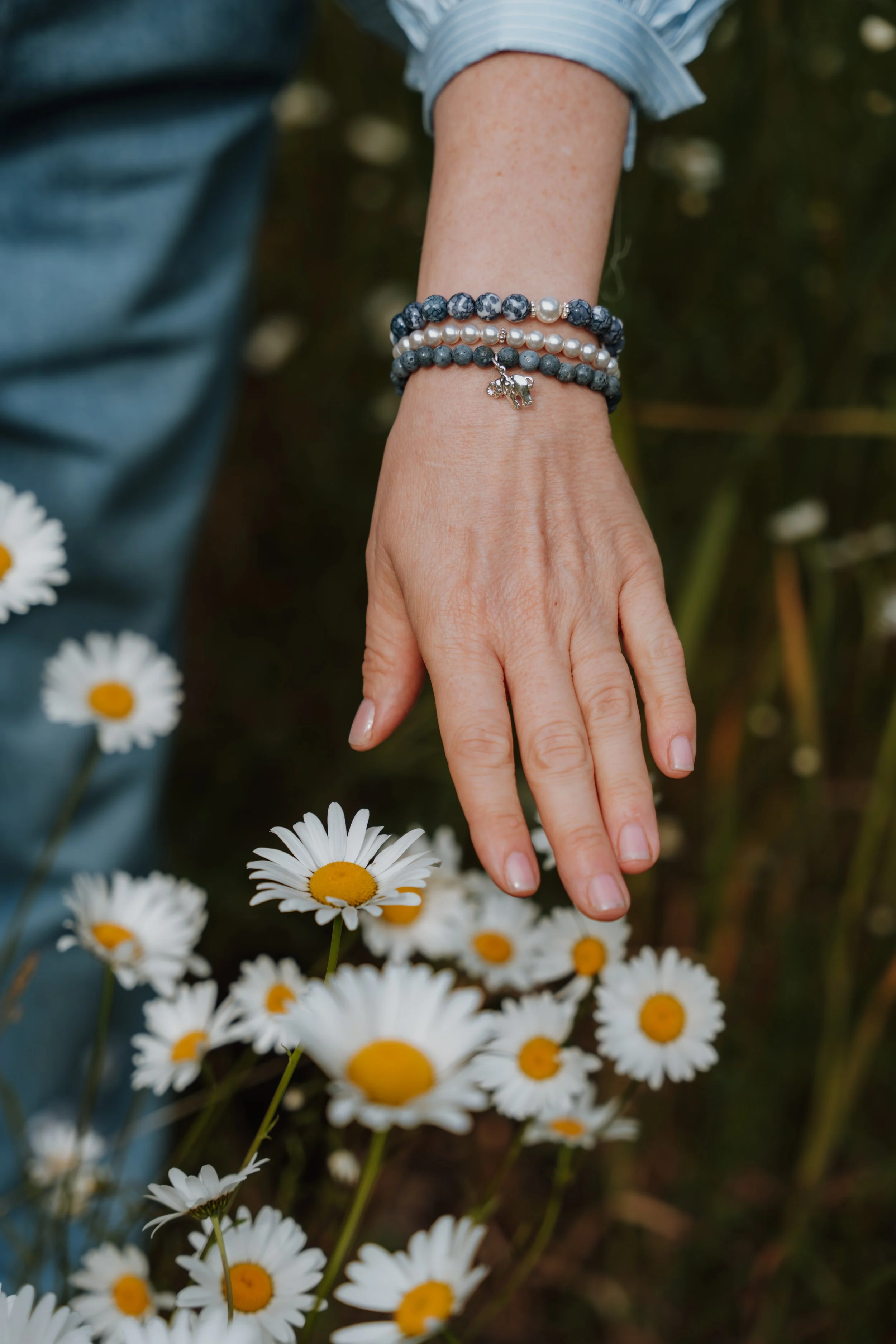 A person's hand and wrist wearing three beaded bracelets, including charms, surrounded by daisies in a natural outdoor setting.