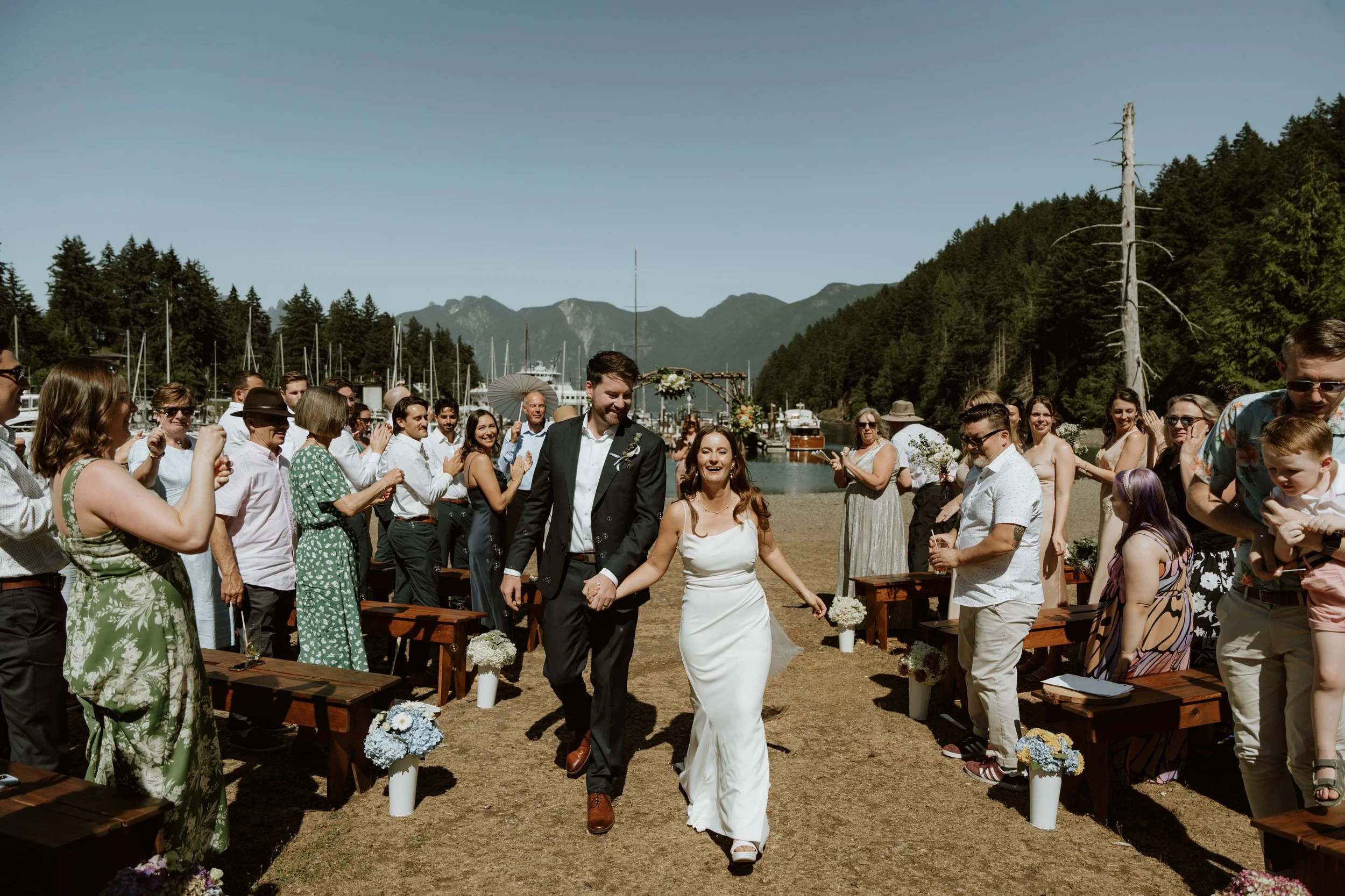 A bride and groom walking down the aisle at an outdoor wedding ceremony near a mountain lake, surrounded by friends and family.