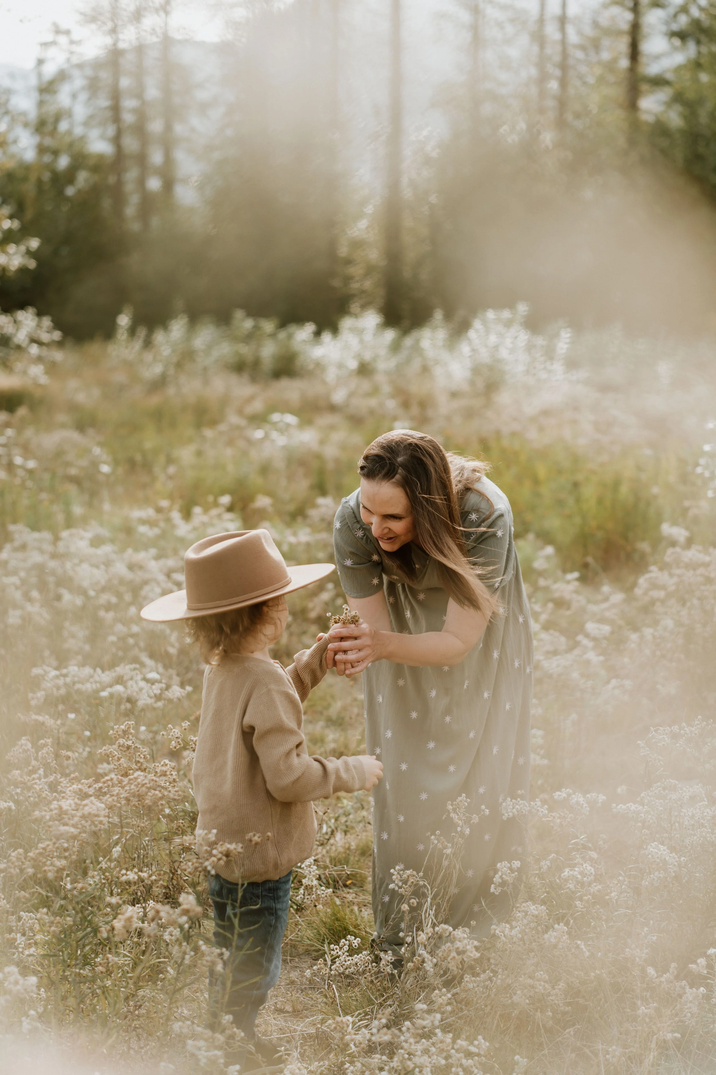 A woman and a young girl sharing a moment in a field of wildflowers during daytime, with the woman offering the girl a small bouquet or flower.