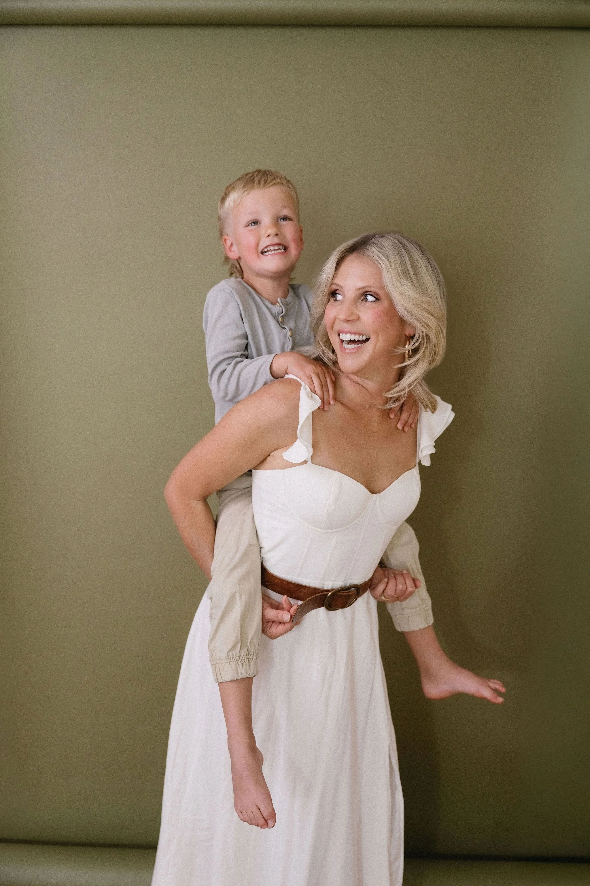 A woman giving a piggyback ride to a young boy with blonde hair, both smiling and laughing against a plain green wall.