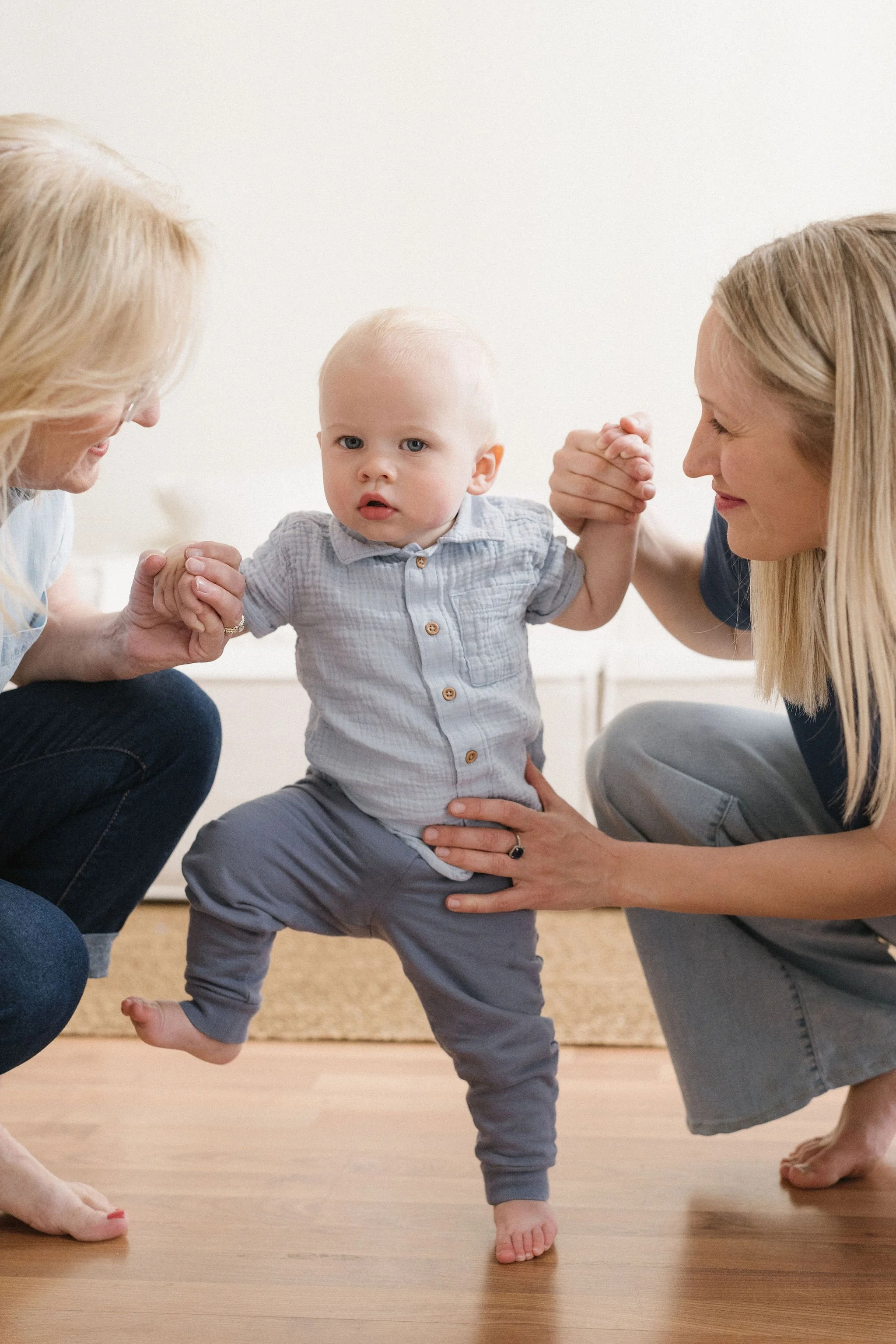 A young child is standing and holding hands with two women, with one woman on each side, while lifting him up gently. The child looks curious and is dressed in a light blue shirt and gray pants. The women are smiling and leaning towards the child, su
