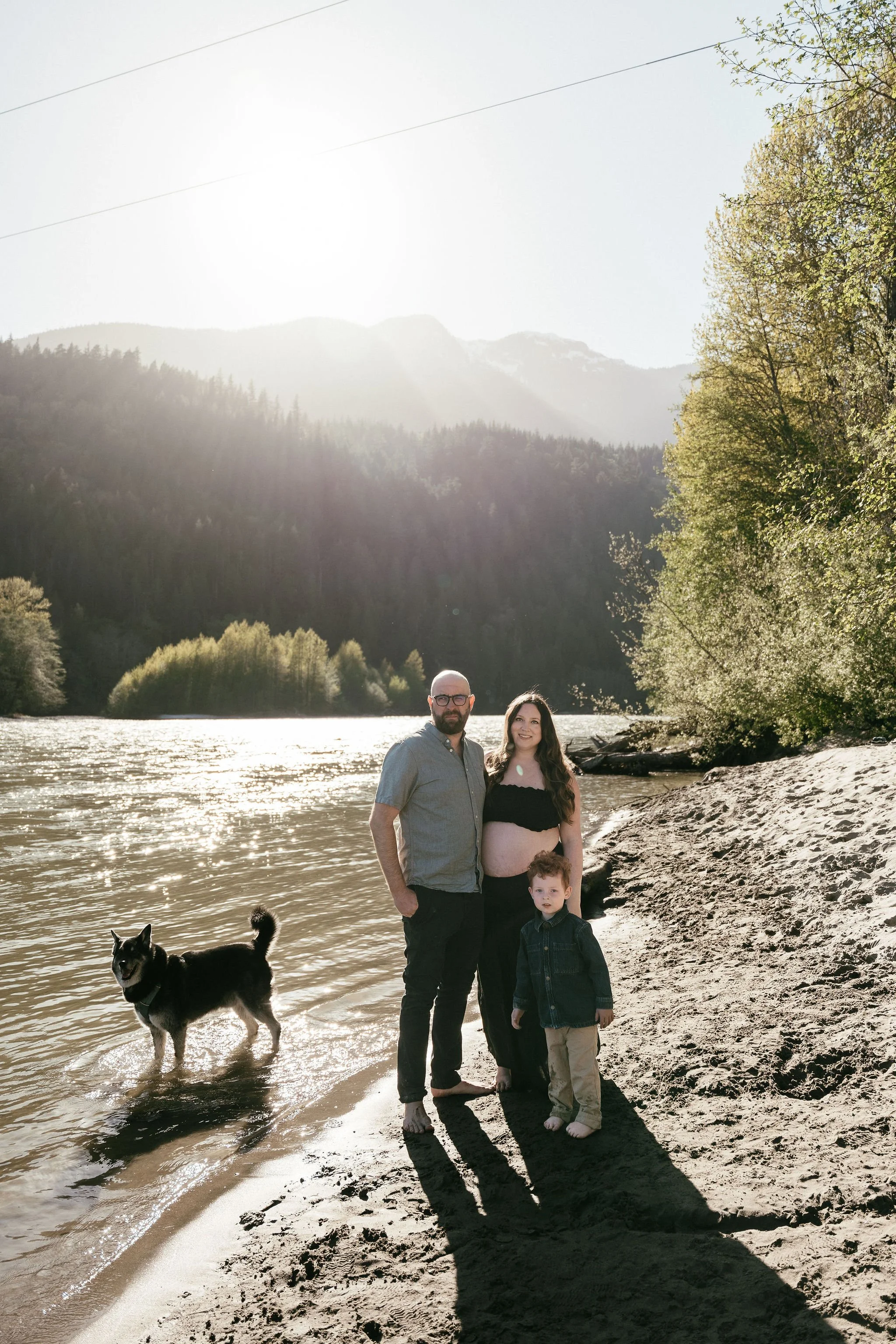 A family of four, including a man, a pregnant woman, a young boy, and a black and white dog, standing on a sandy beach by a river, with green trees, mountains, and a cloudy sky in the background.