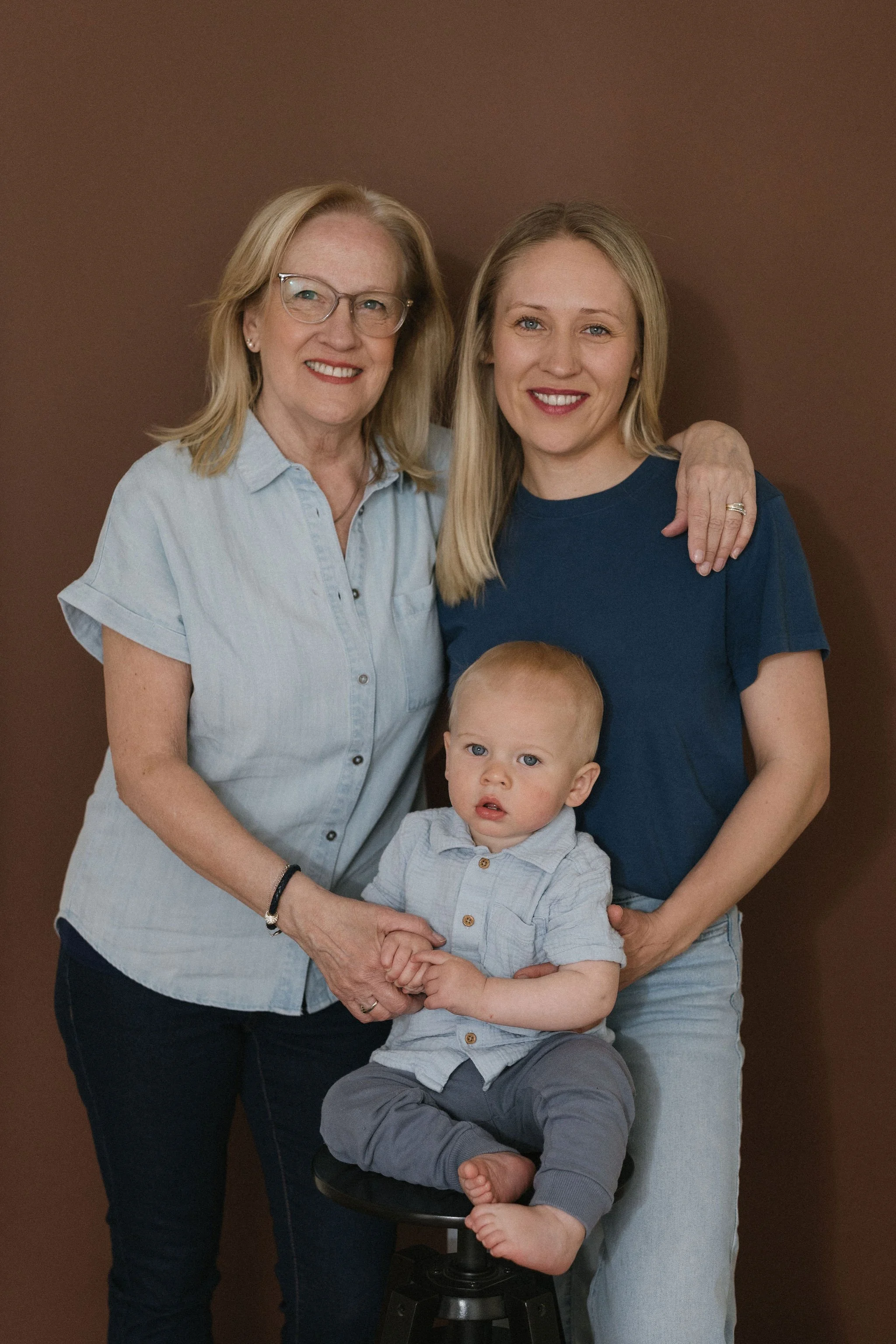 A multigenerational family photo with an elderly woman, a young woman, and a young boy. The elderly woman has blonde hair, glasses, and is wearing a light blue shirt. The young woman has blonde hair and is wearing a navy blue shirt. The boy has blond