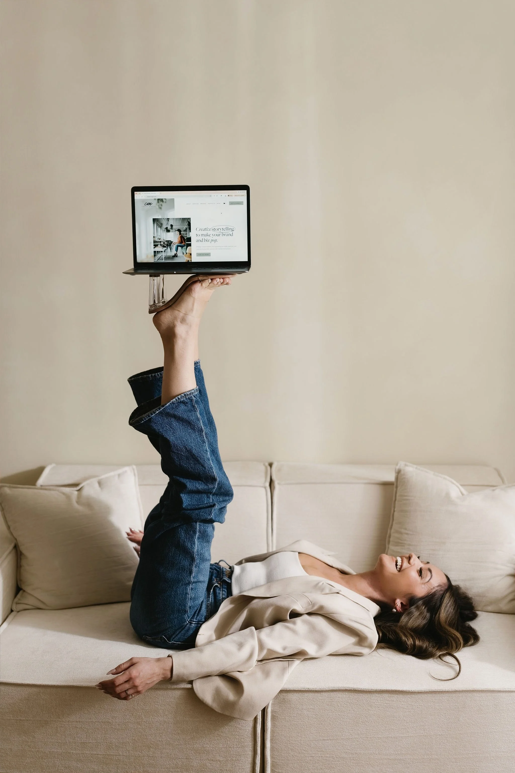 A woman lying on a beige sofa with her head resting on pillows, smiling, while a person holds a small laptop above her with their arm extended. The person’s face is not visible, and they are wearing jeans and a beige jacket. The background features a