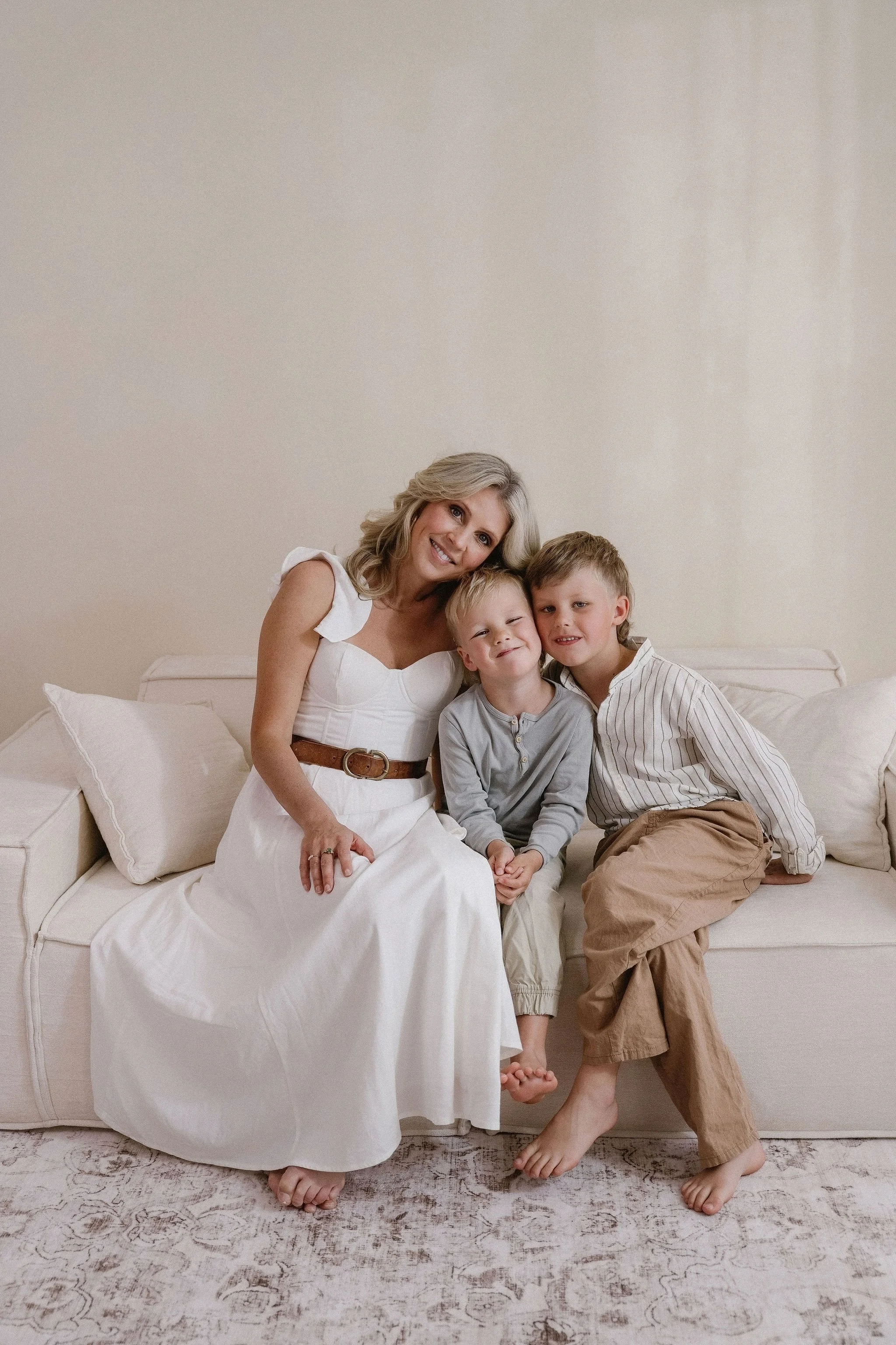 A woman and two young boys sitting on a beige couch in a neutral-colored room. The woman is wearing a white dress and brown belt, smiling, with blond hair styled in loose waves. The boys are casually dressed, barefoot, and sitting close to her, smili