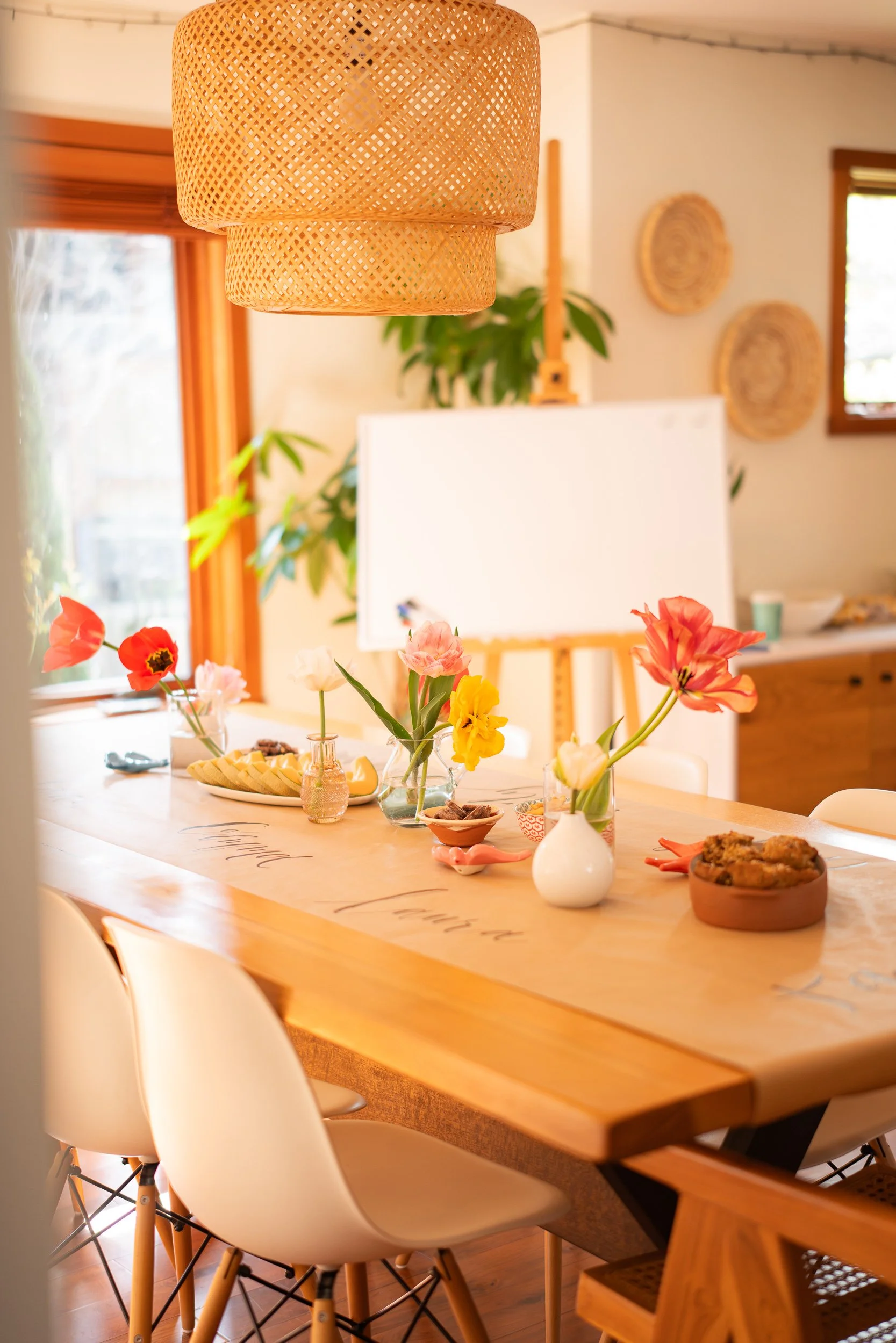 A dining table decorated with colorful flowers in vases, with snacks and pastries, in a bright, cozy room with wooden furniture and woven decorations.