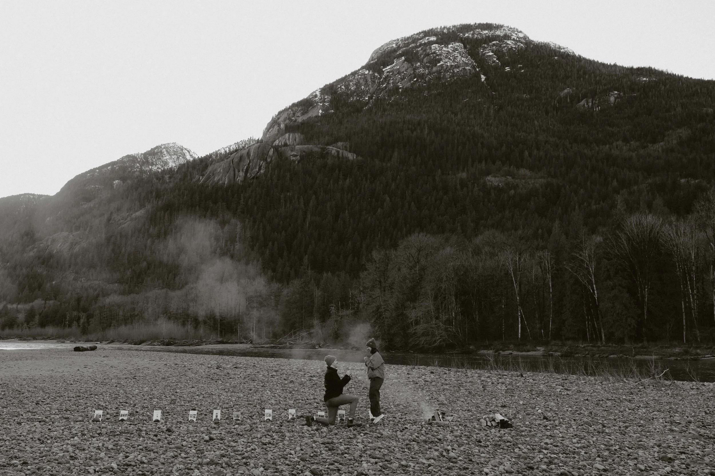 A black and white photo of a mountainous landscape with a forested hillside, a river, and two people on a rocky beach in the foreground. One person is kneeling while the other stands, with small signs or plaques on the ground nearby.