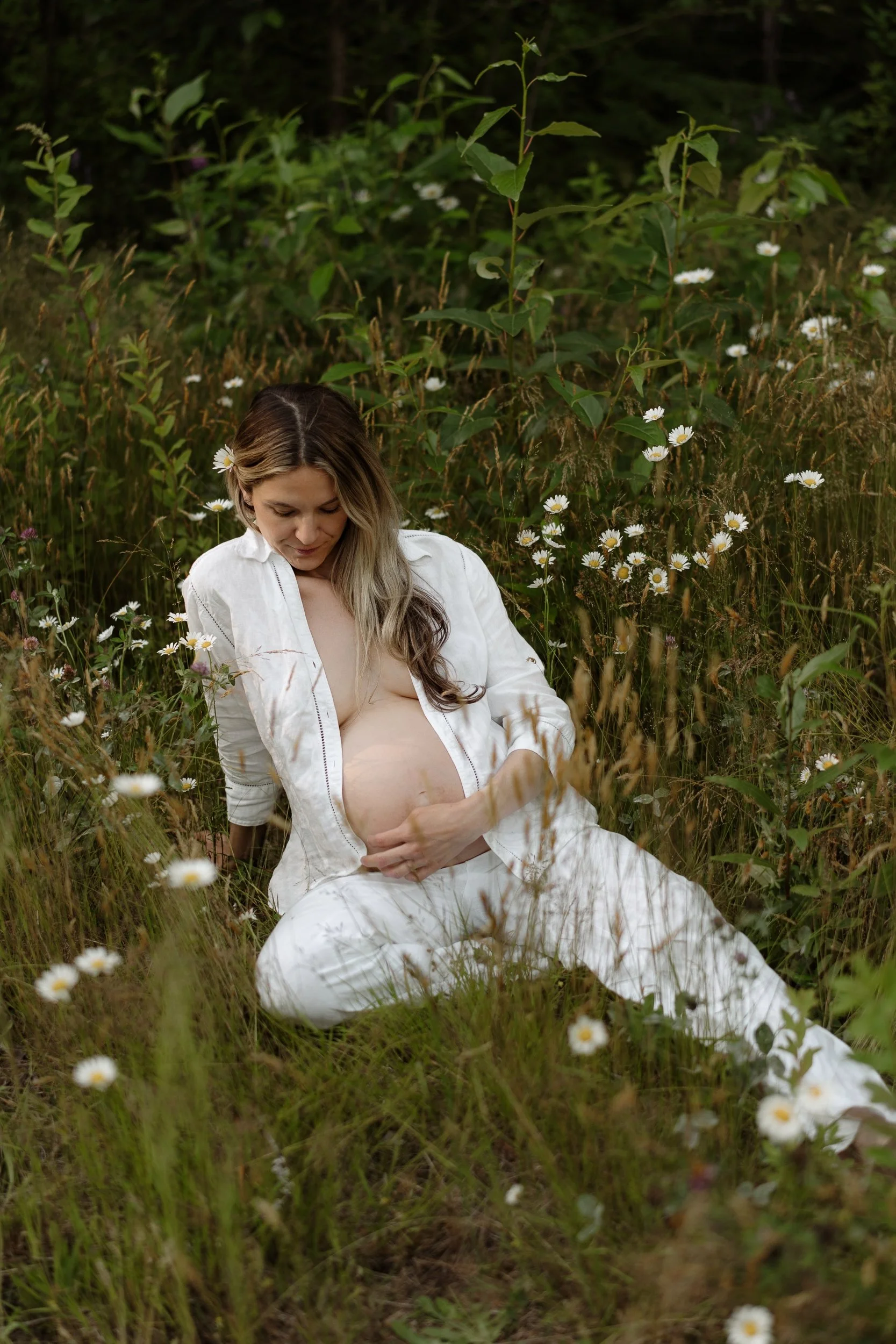 A woman in a white dress sitting in a field of daisies with her hand on her pregnant belly, surrounded by green plants and wildflowers.