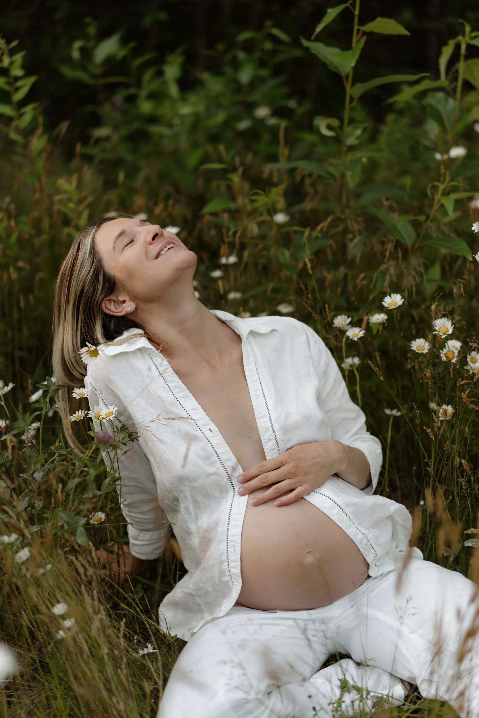 A woman in a white dress lying in a field of daisies and greenery with her eyes closed and a peaceful smile.