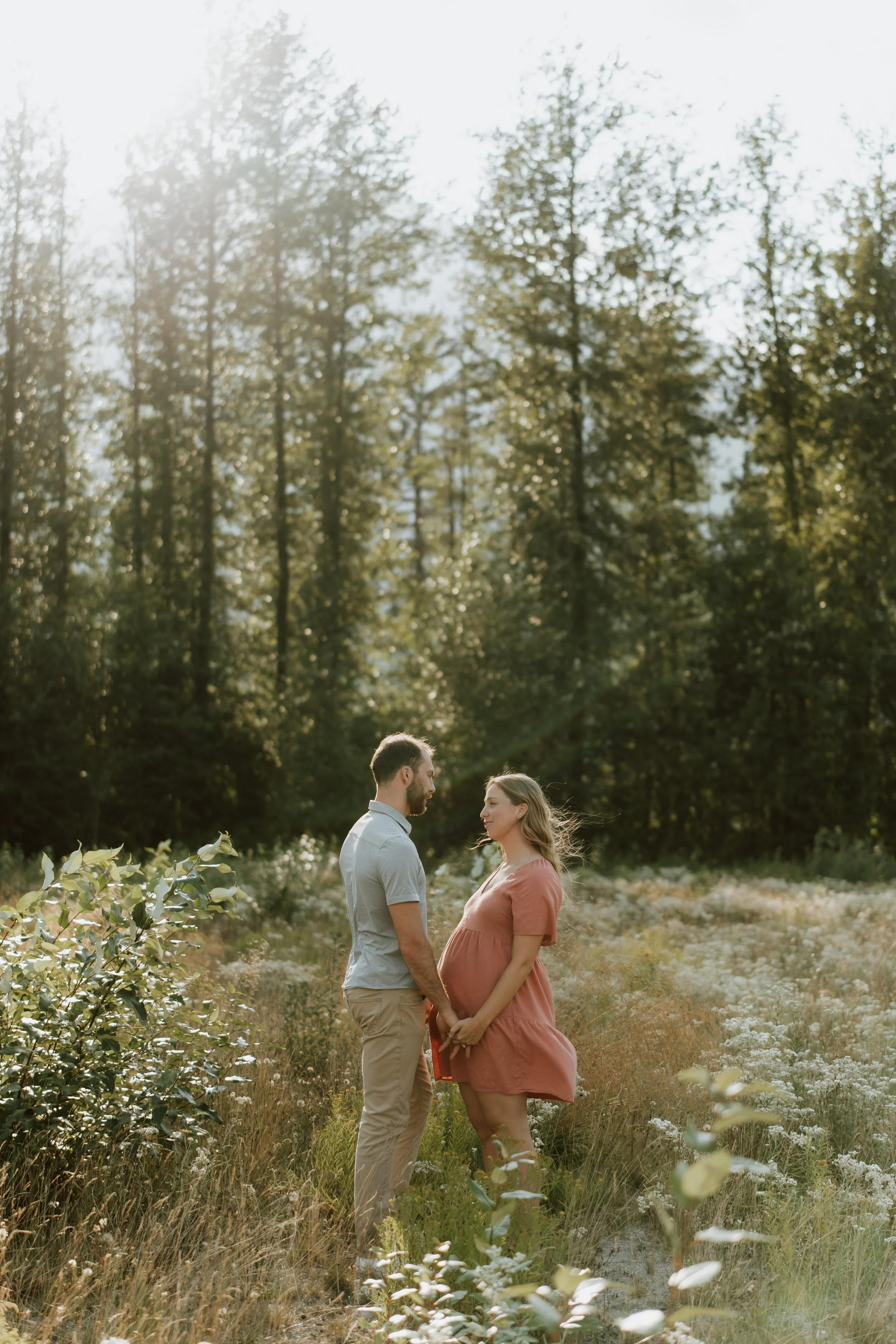 A pregnant woman and her partner standing hand in hand in a field of wildflowers, facing each other with trees in the background and sunlight filtering through the trees.