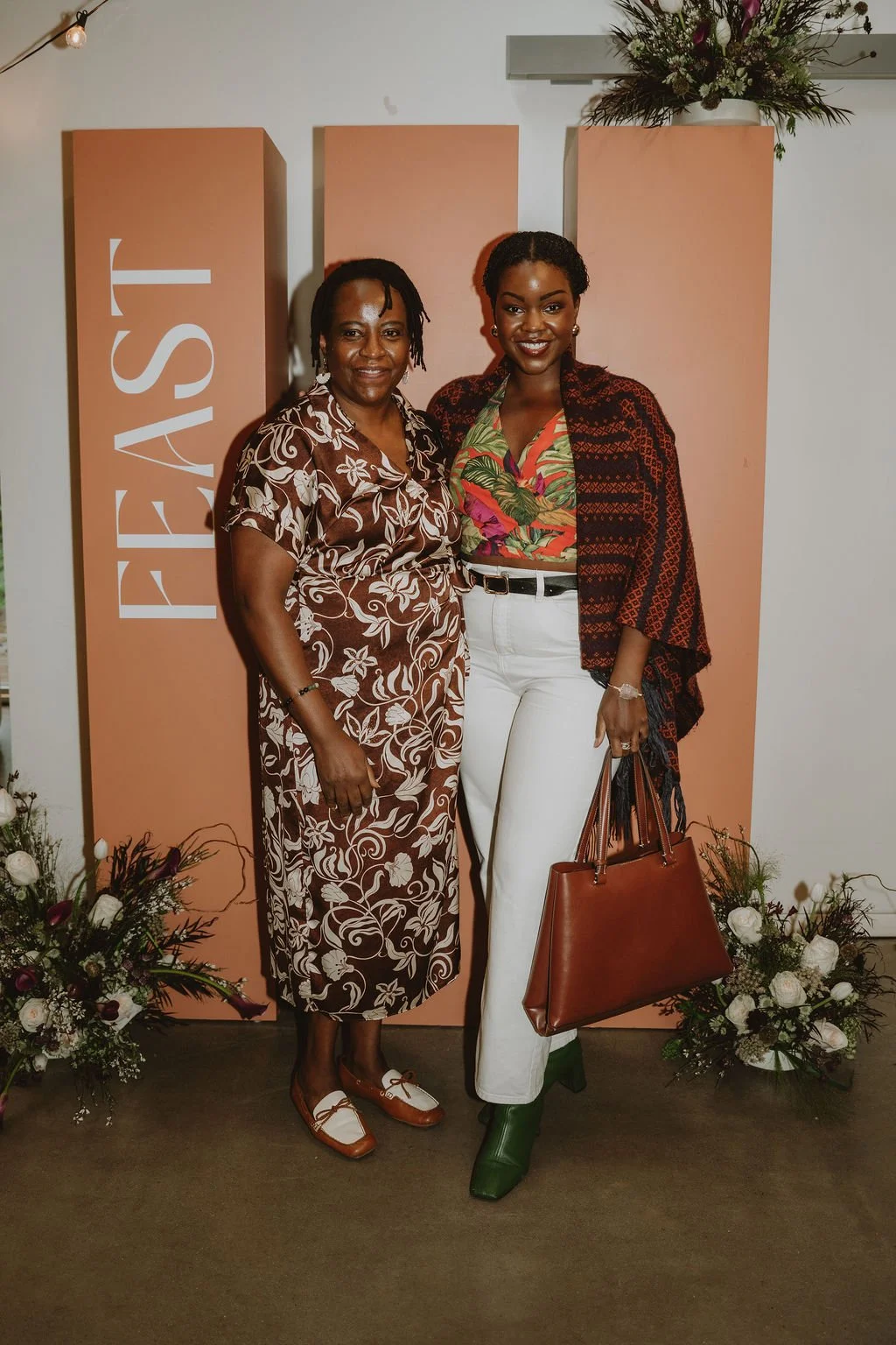 Two women standing together at an event, with a peach-colored backdrop that has the word "FEAST" written vertically on it. They are smiling and dressed in fashionable clothing, surrounded by floral arrangements at their feet.