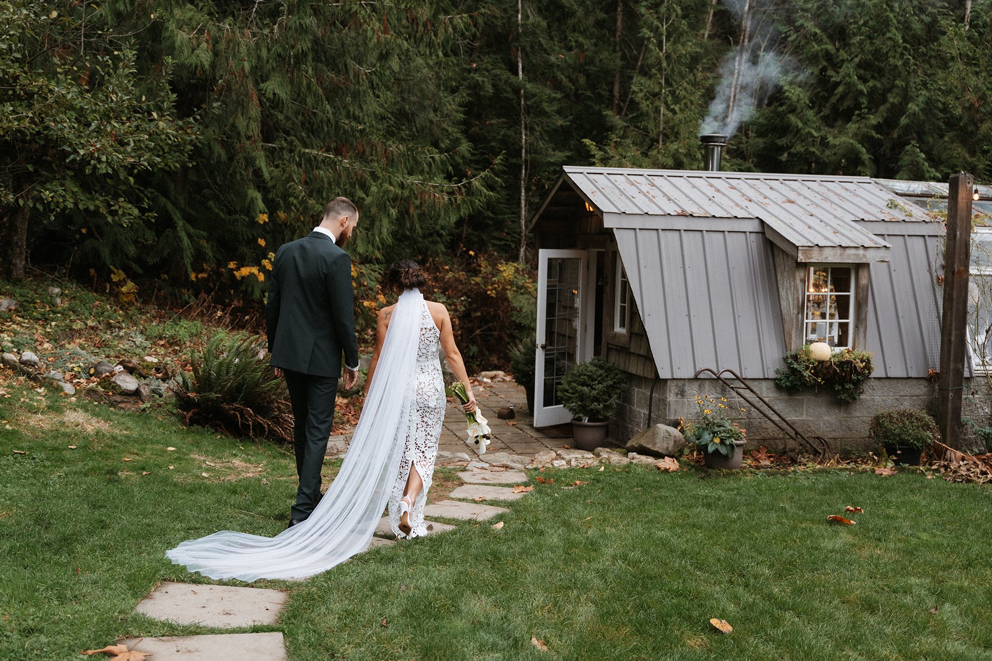 A bride and groom walking on a stone pathway outside in front of a small rustic shed surrounded by greenery. The bride is wearing a white lace dress with a long train and holding a bouquet, and the groom is dressed in a dark suit.