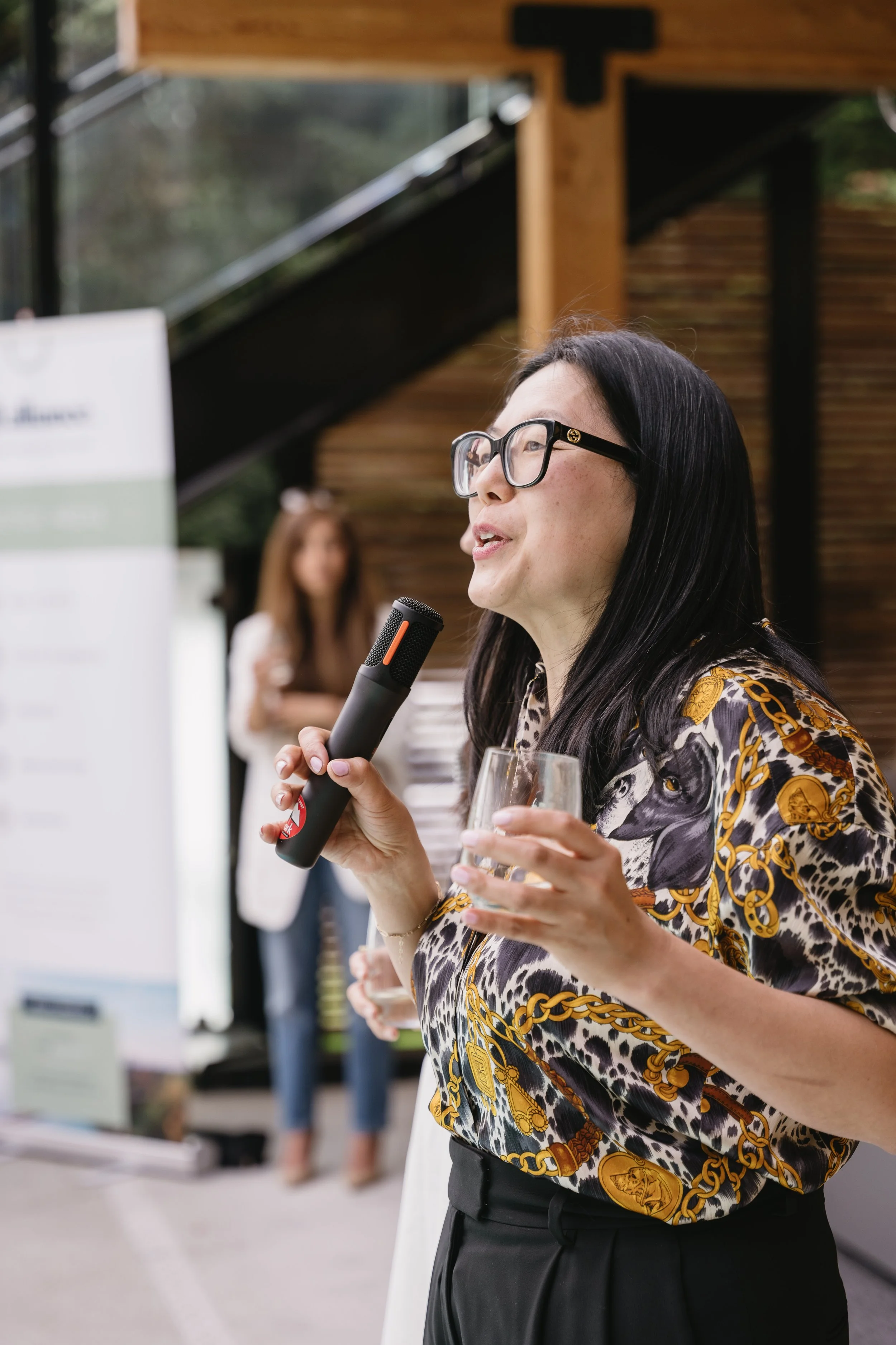 A woman with long black hair, glasses, and a leopard and gold chain patterned blouse speaking into a microphone at an outdoor event, holding a glass of champagne.