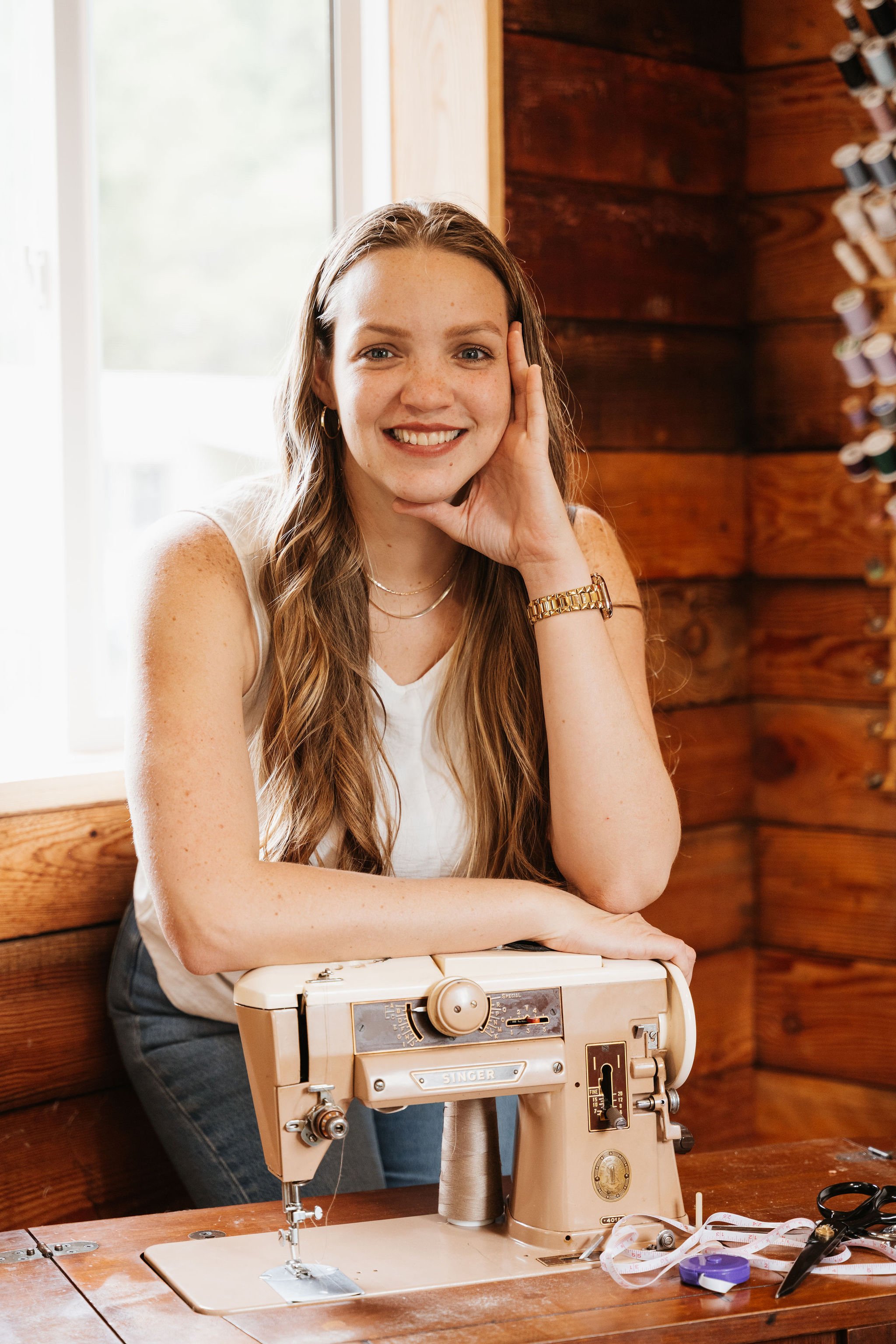 A woman sitting at a wooden table with a vintage sewing machine, smiling, in a cozy, rustic room with wooden walls and a window.