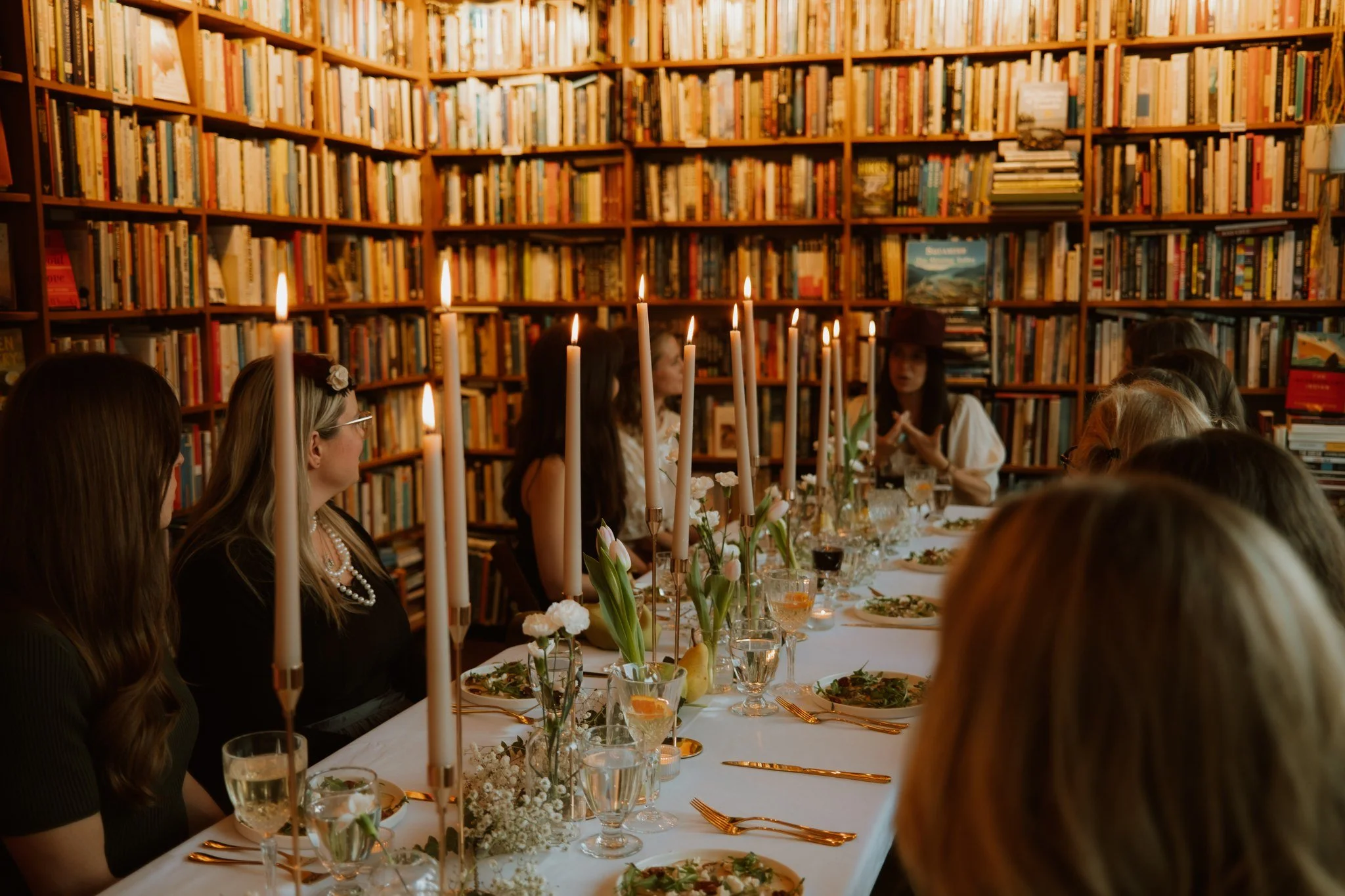 People sitting around a dinner table with tall candles and floral centerpieces, inside a room with bookshelves filled with books.