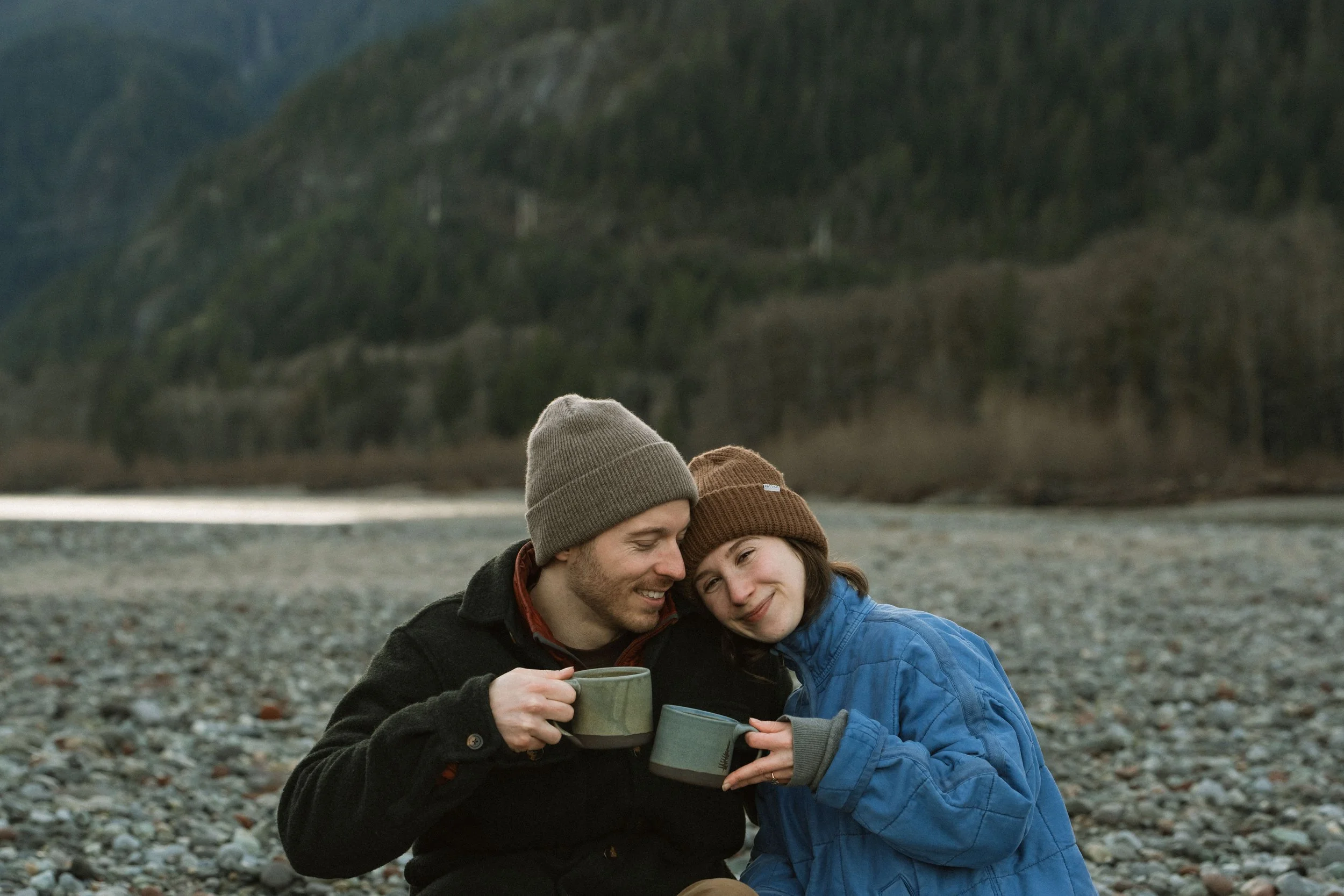 A man and a young girl sitting close together on a rocky beach, holding mugs and smiling with their eyes closed, wearing warm clothing and beanie hats.