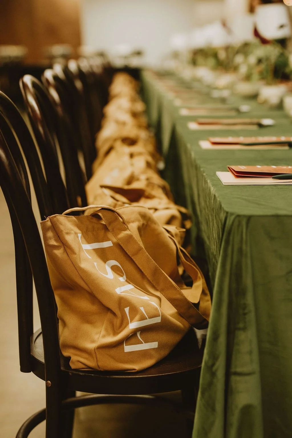 A beige tote bag with the word 'YES' printed on it in white, placed on a black chair at a banquet table with green tablecloths, set with papers and utensils.