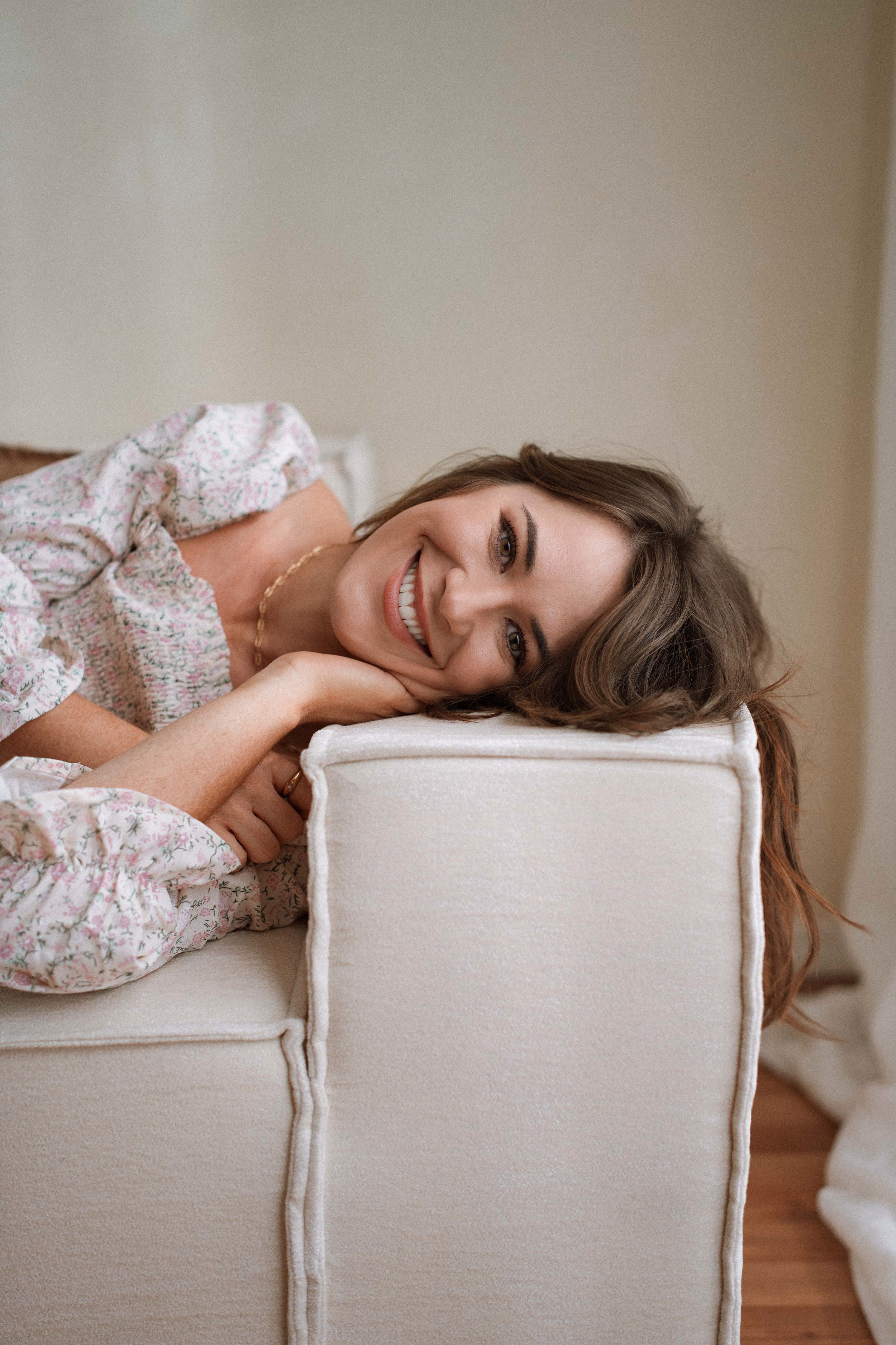 A smiling woman with brown hair lying on a white sofa, resting her head on a pillow with a floral pattern, wearing a floral dress and a necklace.