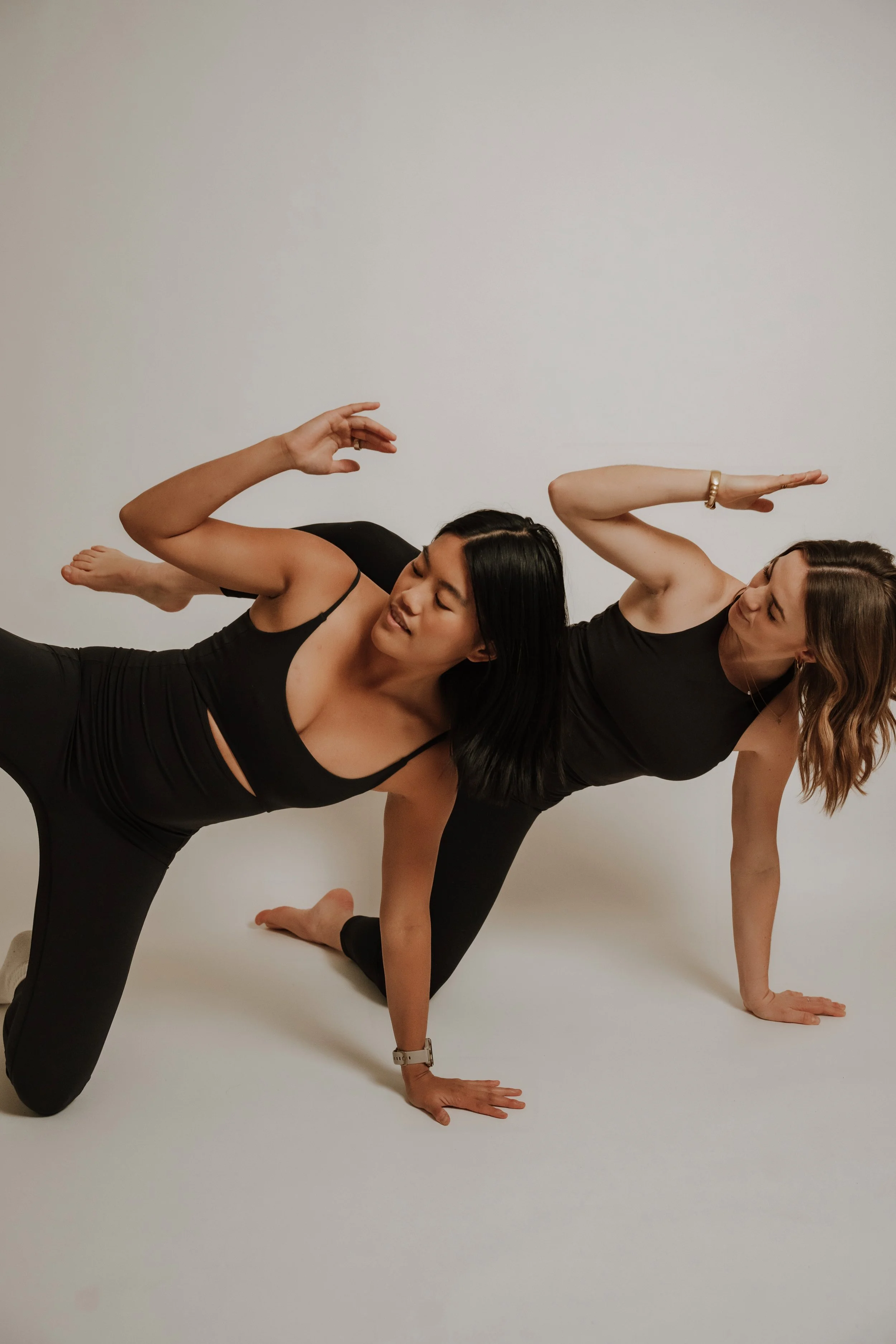 Two women practicing yoga on the floor, performing side plank poses with their right hands on the ground, wearing black athletic wear, against a plain white background.