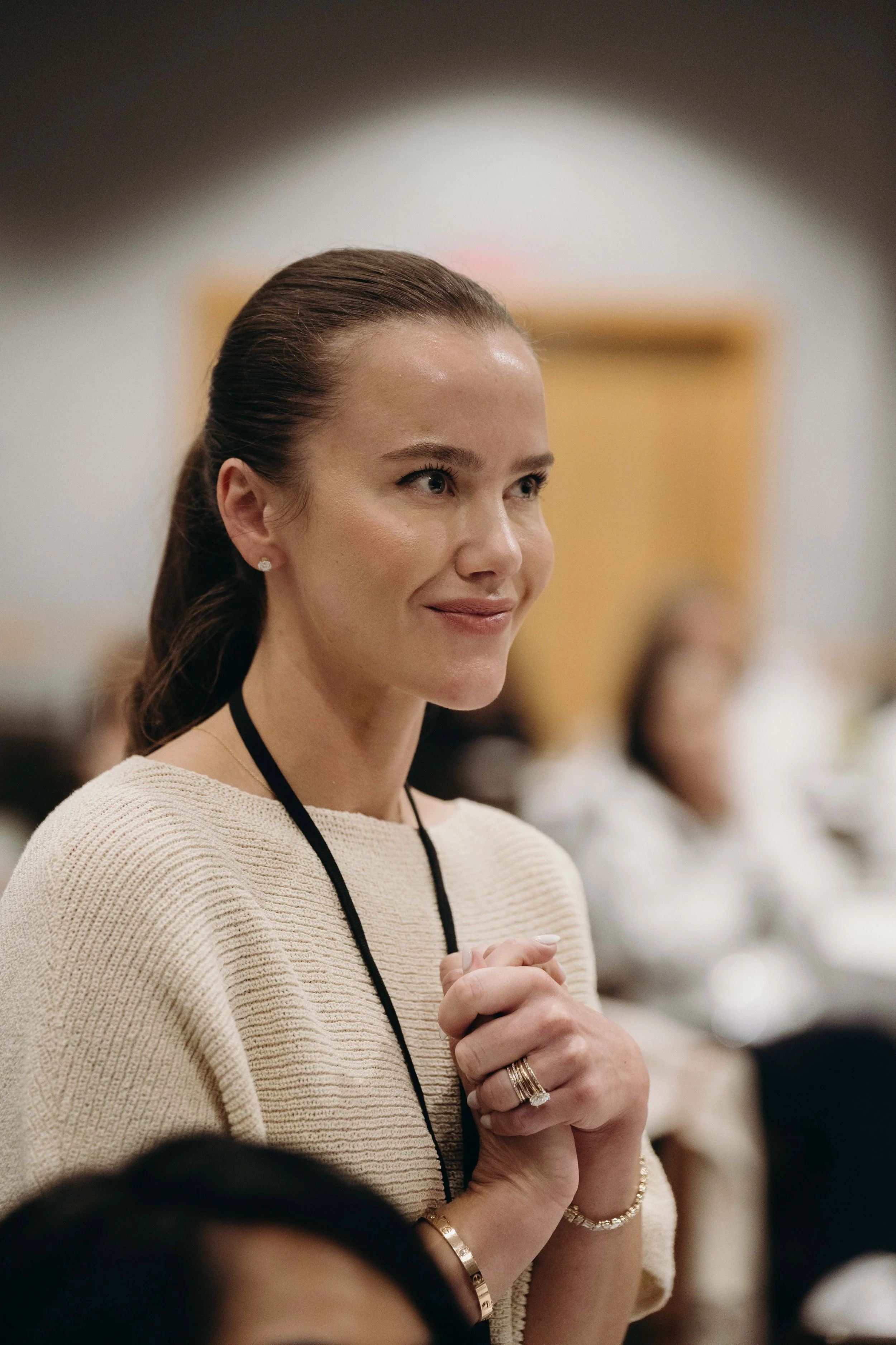 A woman with brown hair styled in a ponytail, wearing a cream-colored sweater, earrings, and jewelry, is standing indoors with her hands clasped, smiling softly, during a formal event or conference.