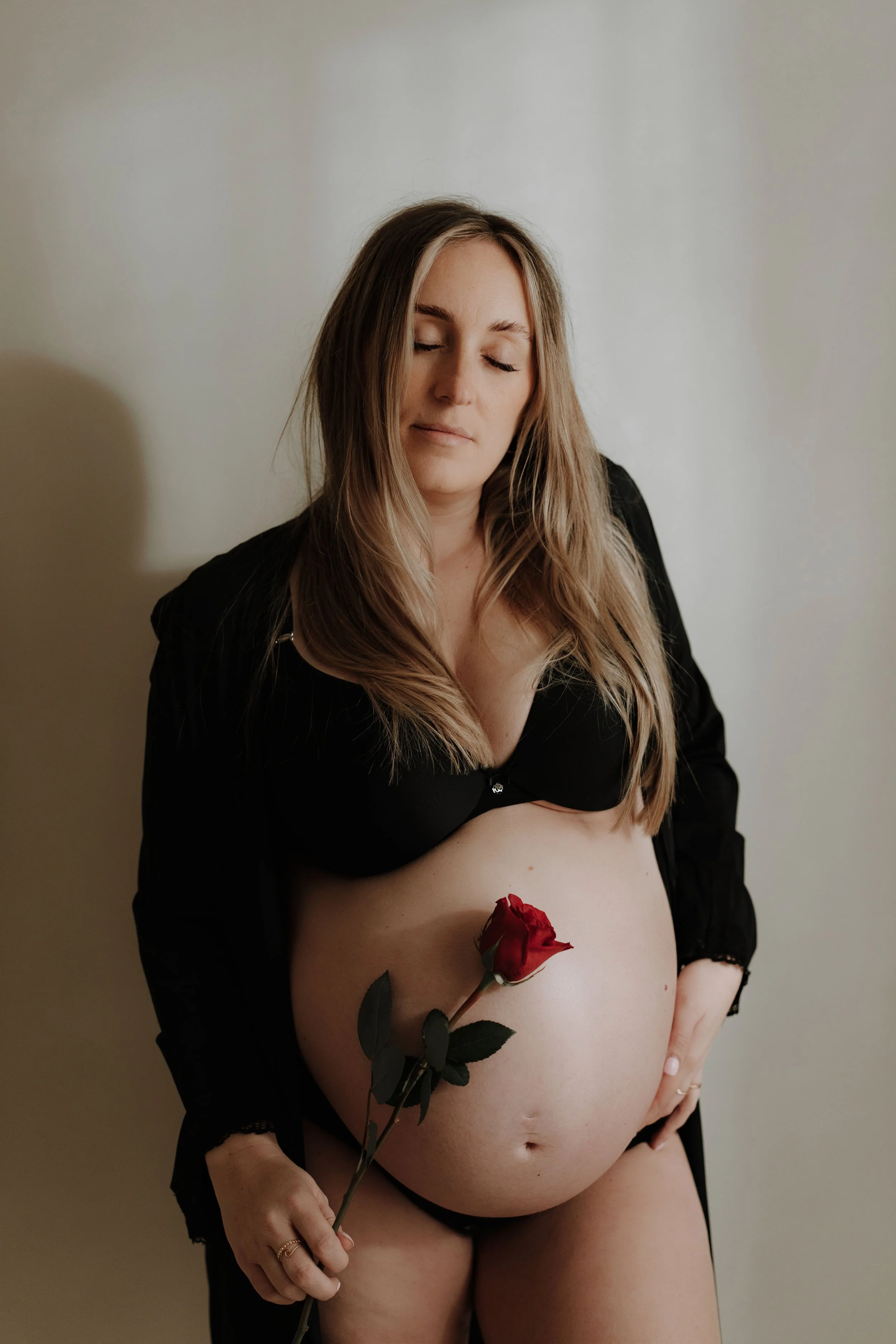 A woman with long, wavy hair and closed eyes, wearing a black robe, holding a red rose, with her midsection exposed and resting her hand on her hip.
