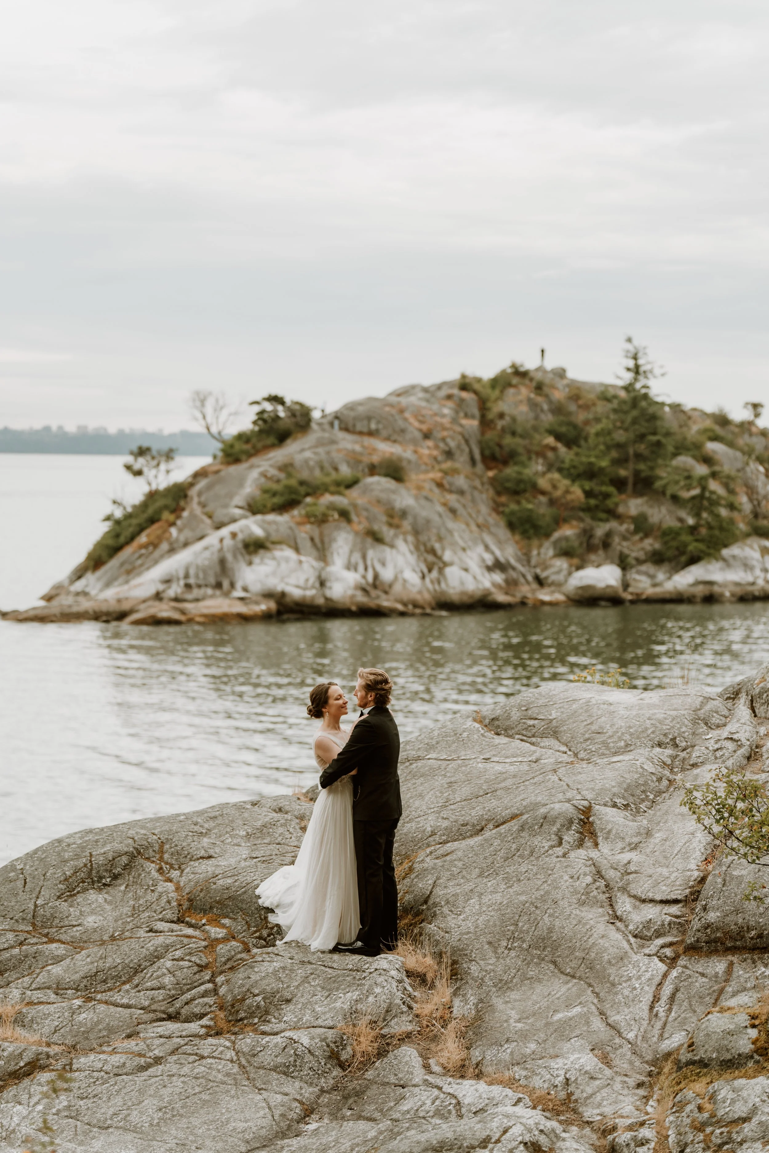 A bride and groom standing on large rocks by a lake, embracing with a scenic mountainous background and cloudy sky.