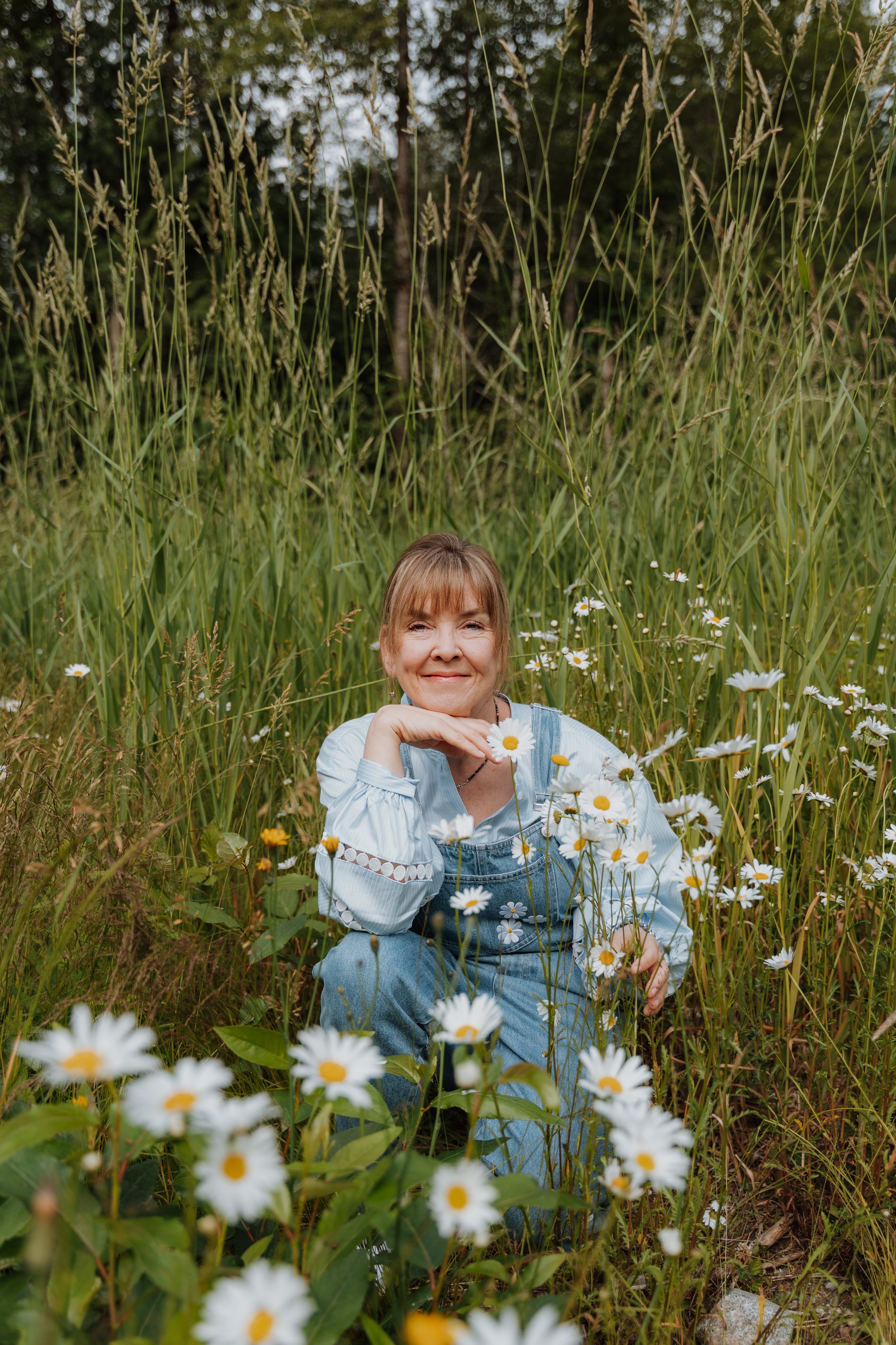 Woman smiling and sitting in a field of daisies with tall grass and trees in the background.