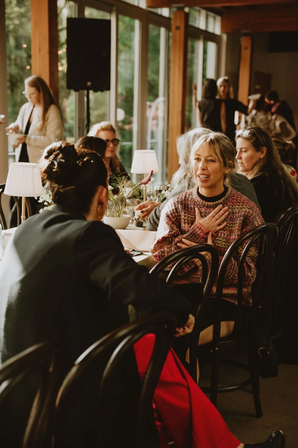 A woman with blond hair, wearing a patterned sweater, gestures with her hand on her chest and appears to be speaking or explaining something during a gathering inside a warmly lit room with large windows. Other women are seated around the table, and 