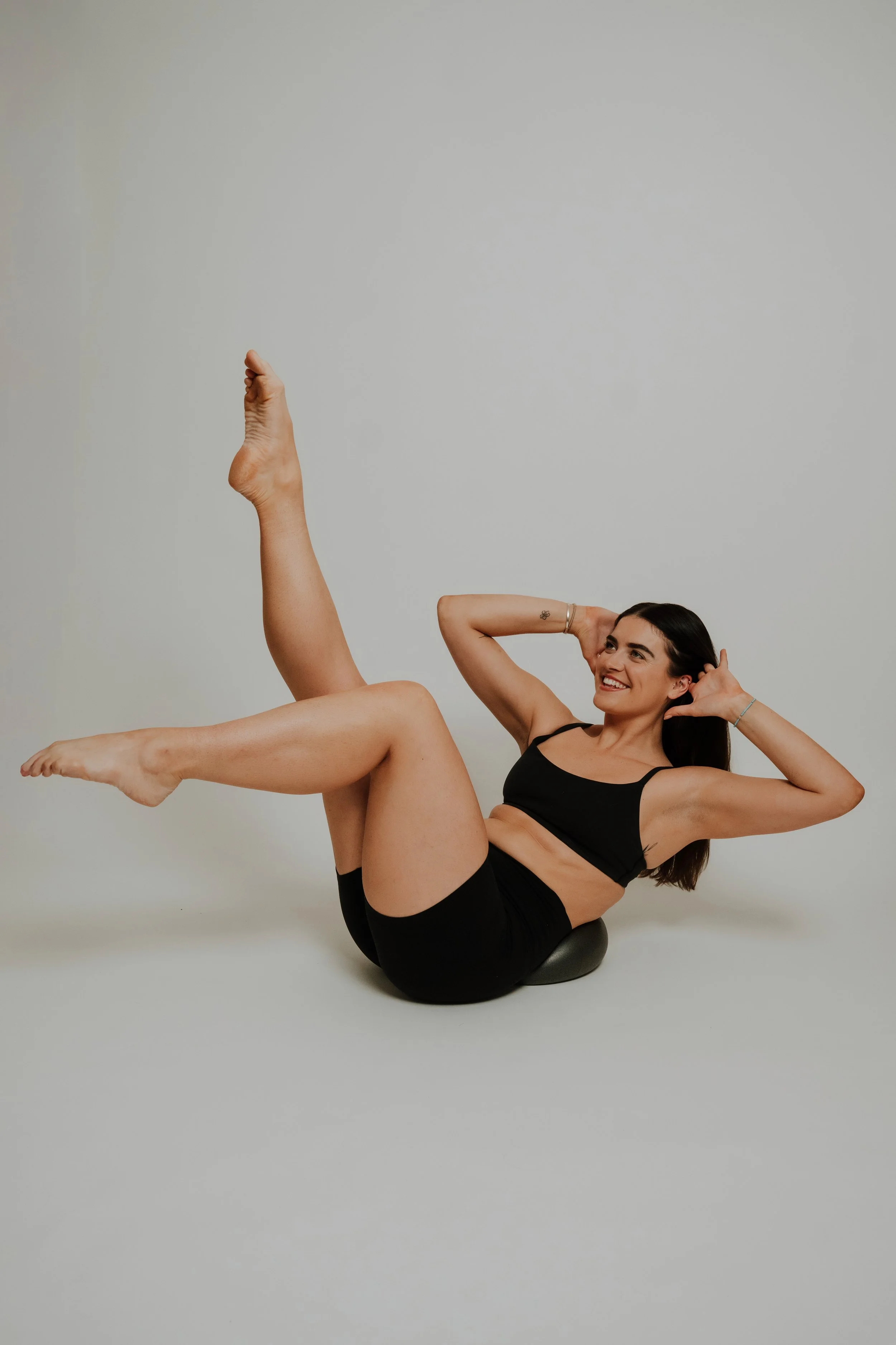 A woman doing a yoga pose on a black ball, lying on her back with one leg raised and her hands behind her head, smiling.