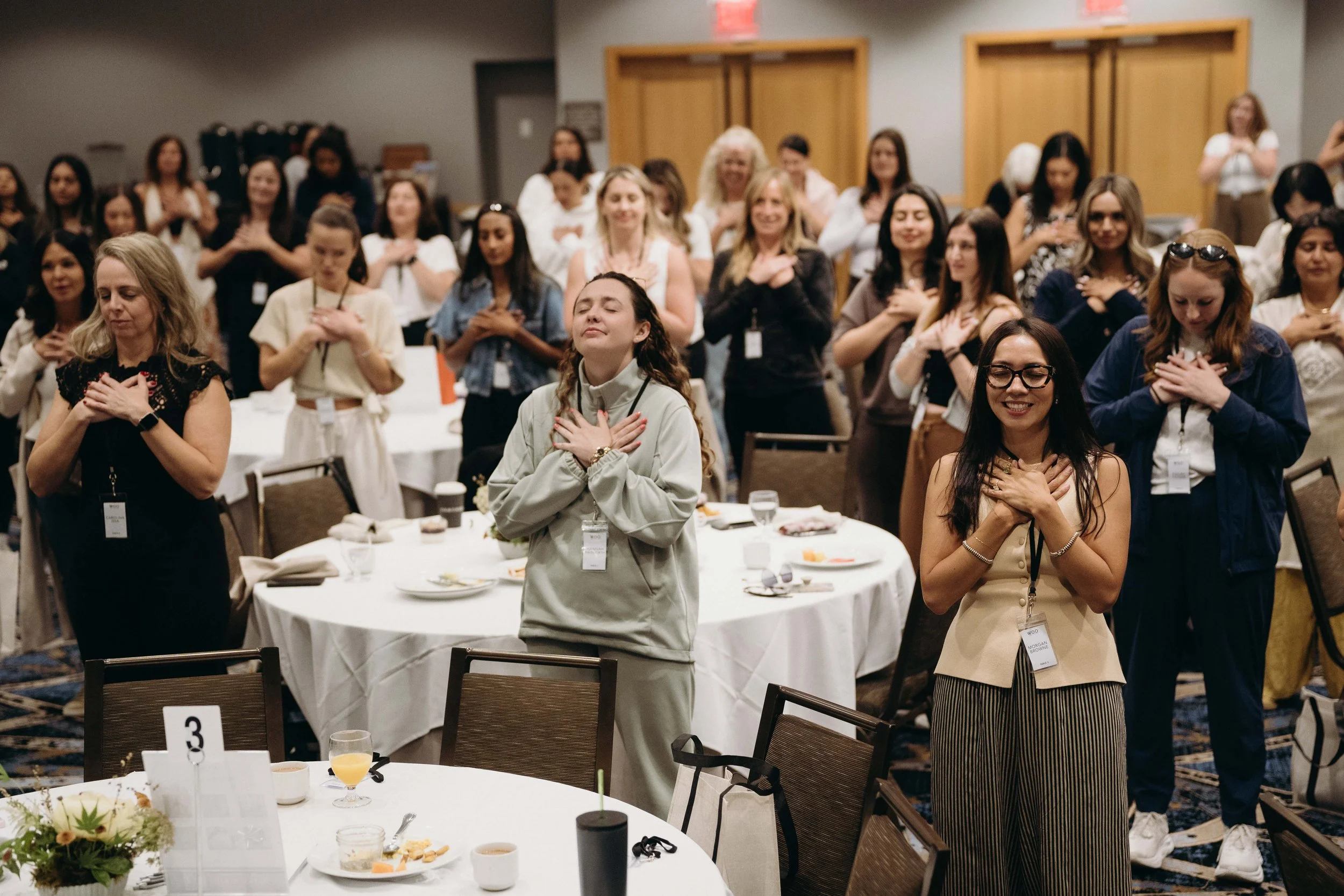 A group of women standing with hands over their hearts during a ceremony at a conference or event, with smiling faces and some wearing name tags.