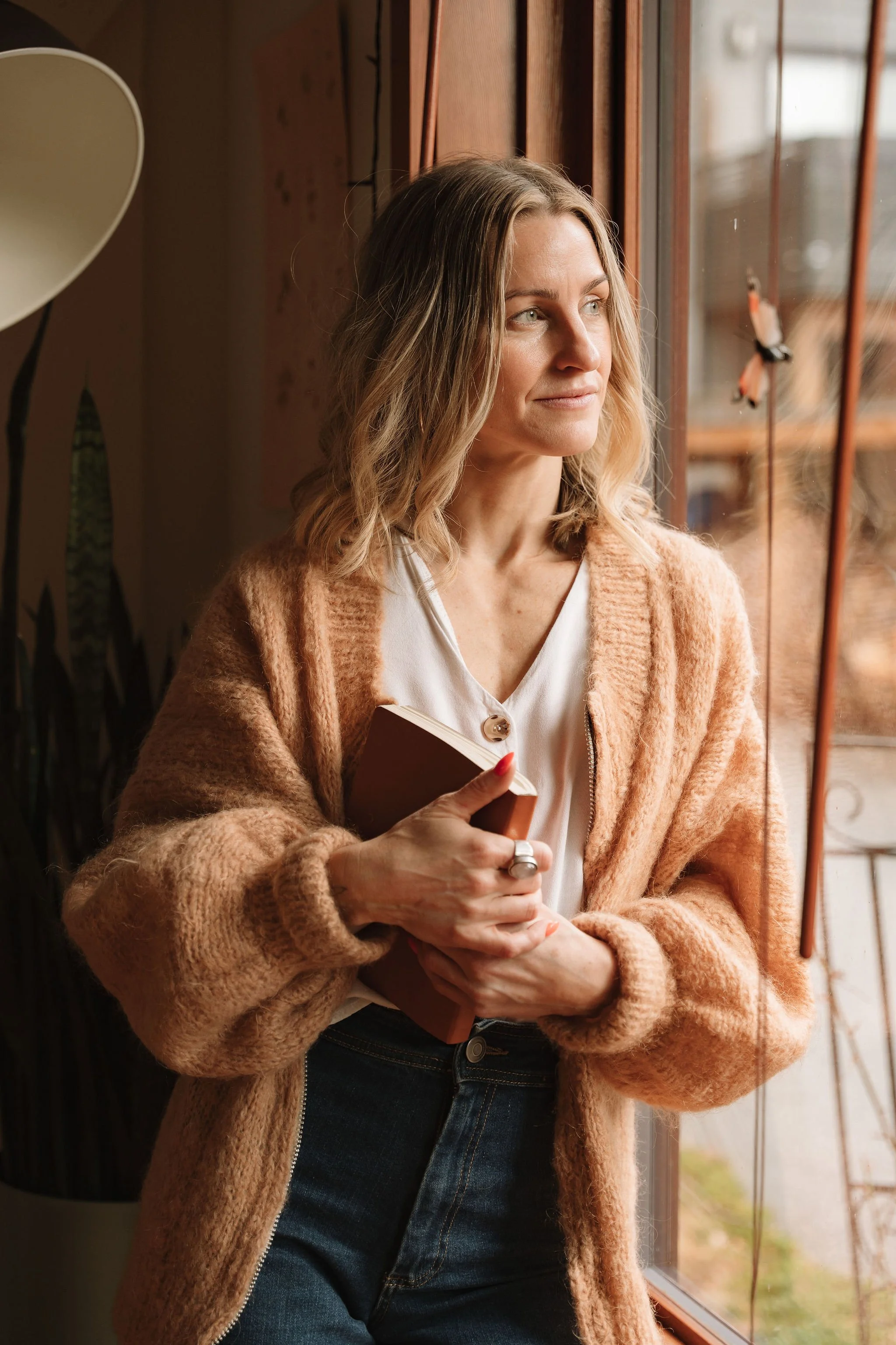 Woman with wavy hair looking out of a window, holding a notebook, wearing a tan cardigan and dark jeans.