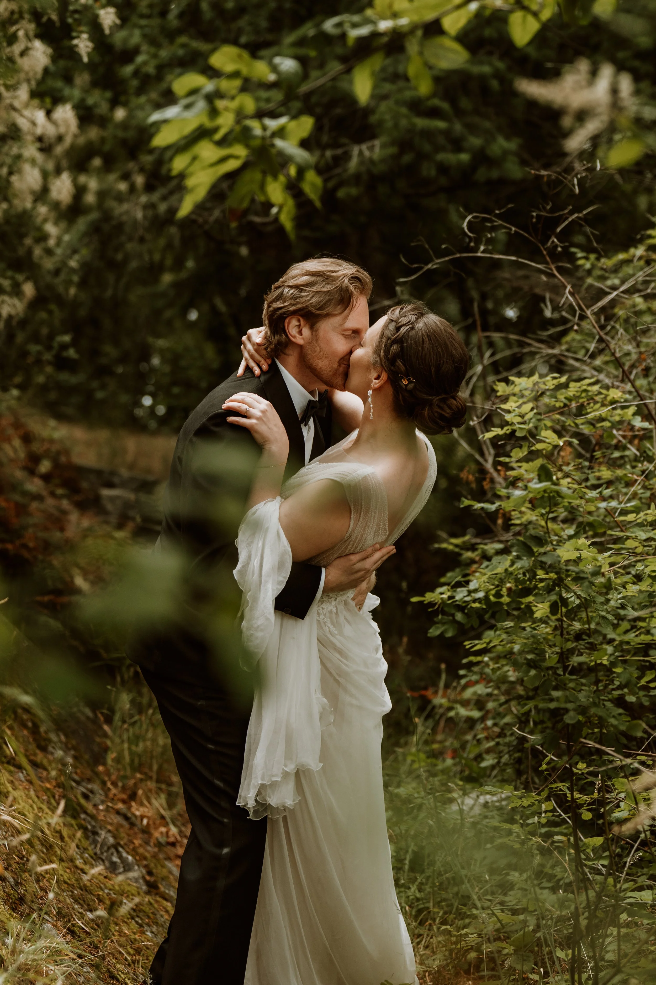 A couple in wedding attire sharing a kiss in a lush, green outdoor setting surrounded by trees and foliage.