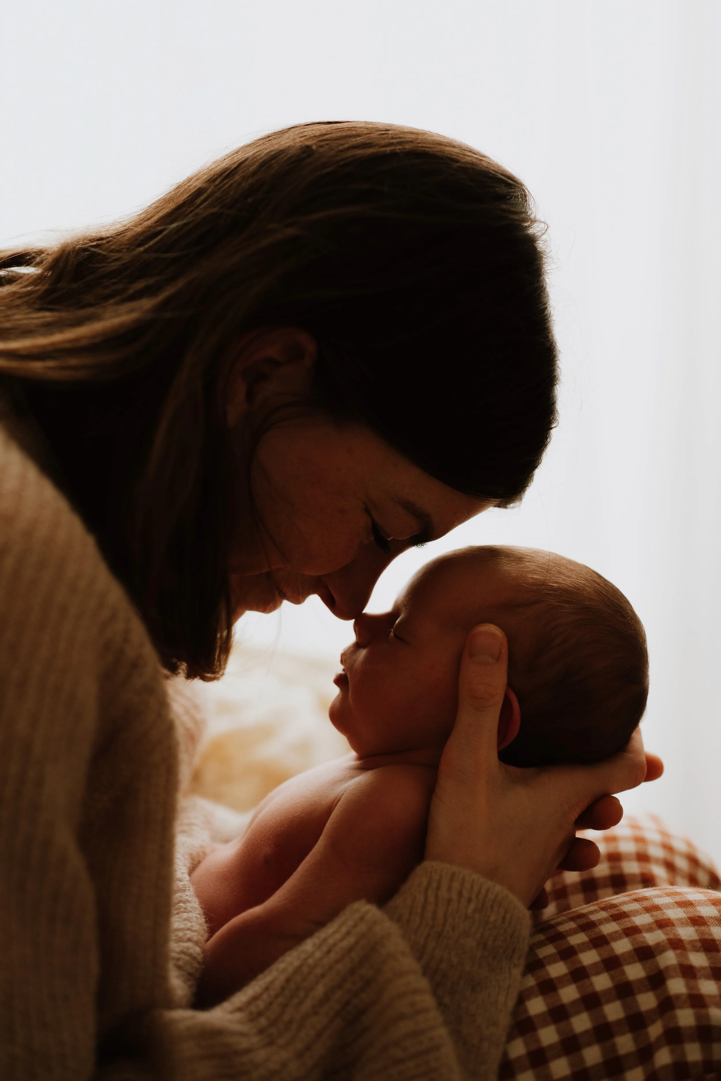 A woman with brown hair holding a sleeping baby close to her face, gently touching the baby's forehead.