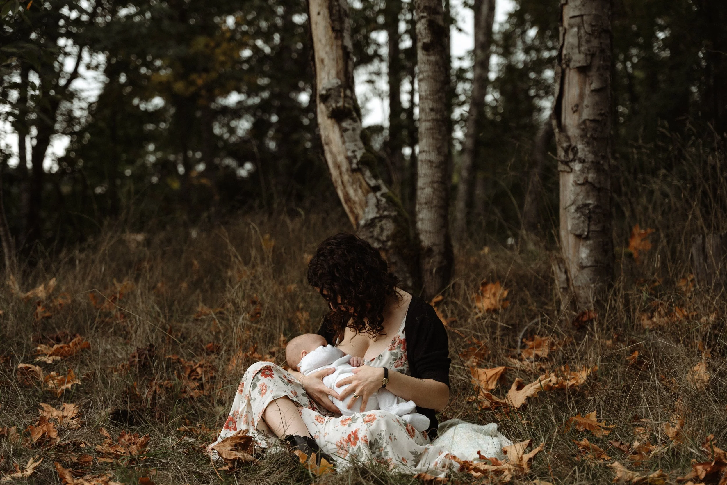 A woman with curly hair, wearing a floral dress and black cardigan, sitting on the ground in a wooded area, breastfeeding a baby wrapped in a white blanket. There are autumn leaves scattered on the ground and trees in the background.