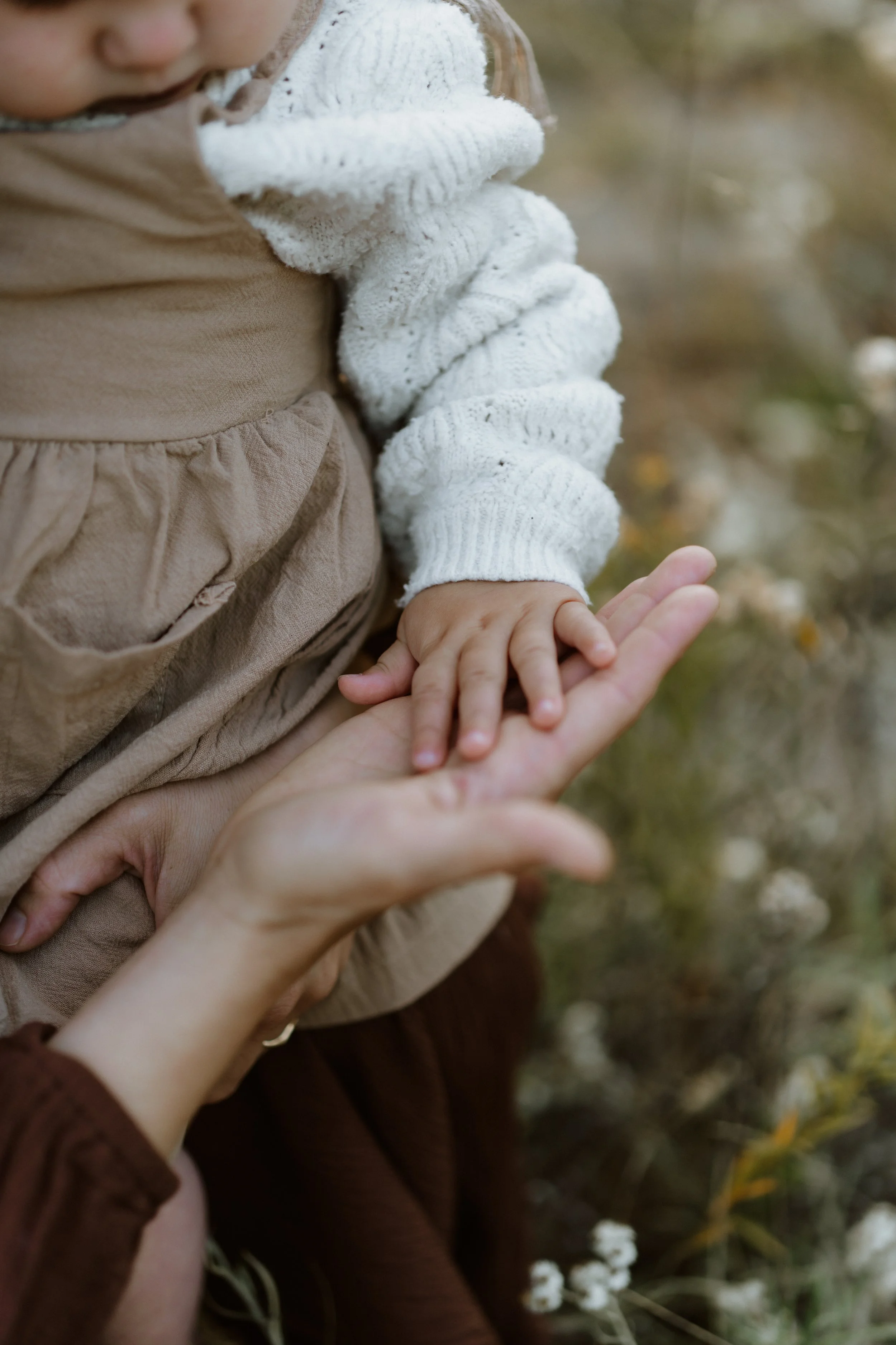 Close-up of a child's hand touching an adult's hand outdoors, with blurred background of plants and flowers.