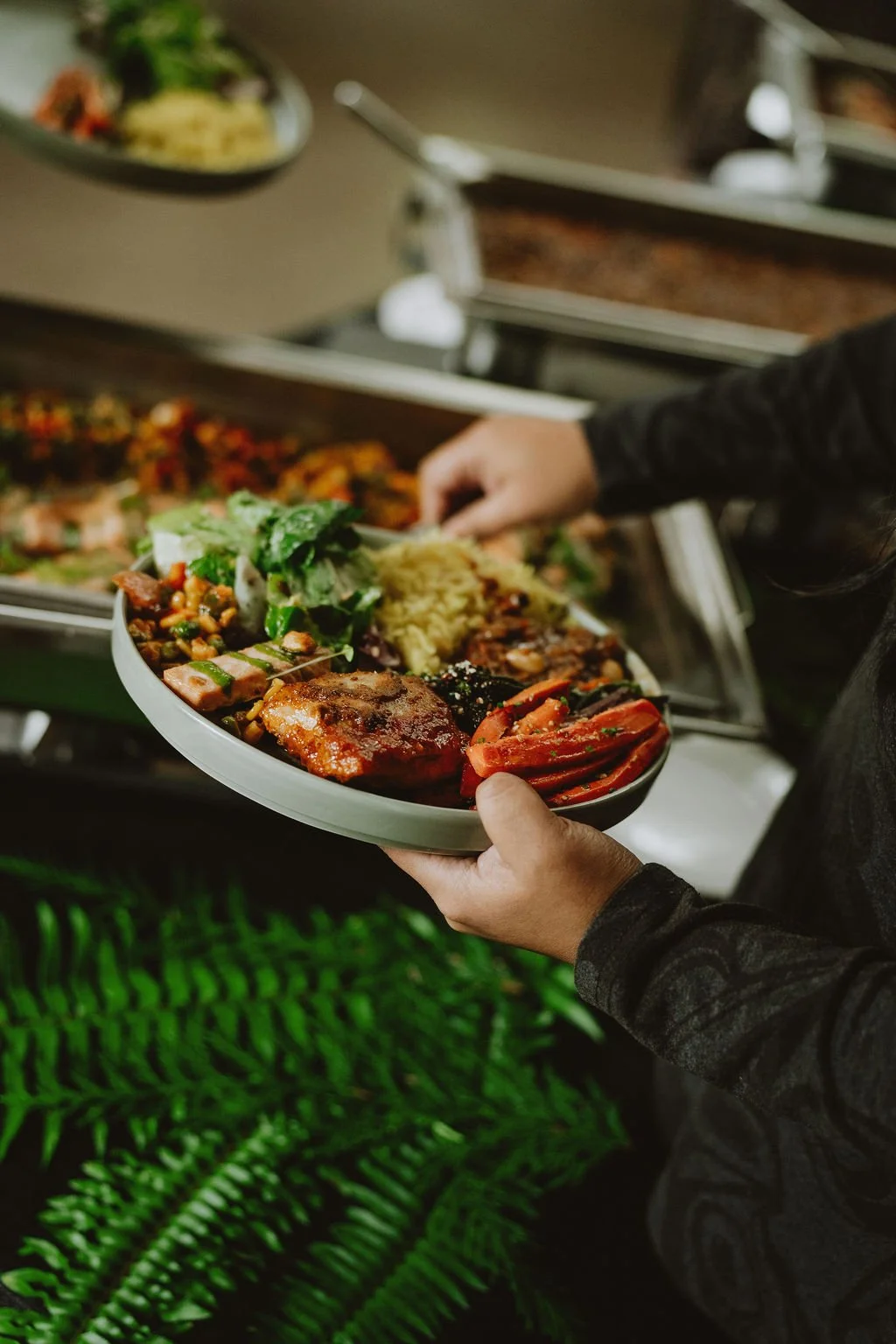 Person serving themselves food from a buffet with a plate of various dishes, including tomatoes, greens, and grilled items, with potted plants in the background.
