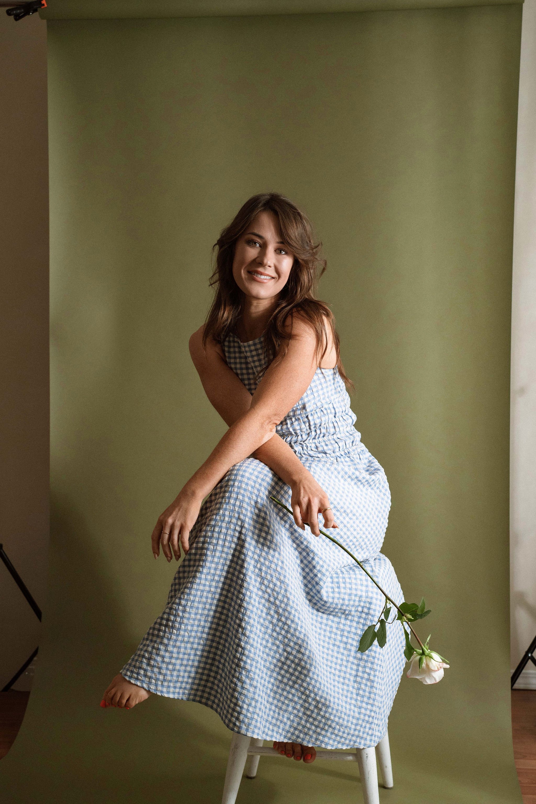 A woman with brown hair, wearing a blue and white checkered dress, is sitting on a stool holding a rose with green leaves in her hand. She is smiling and posed against a green backdrop.