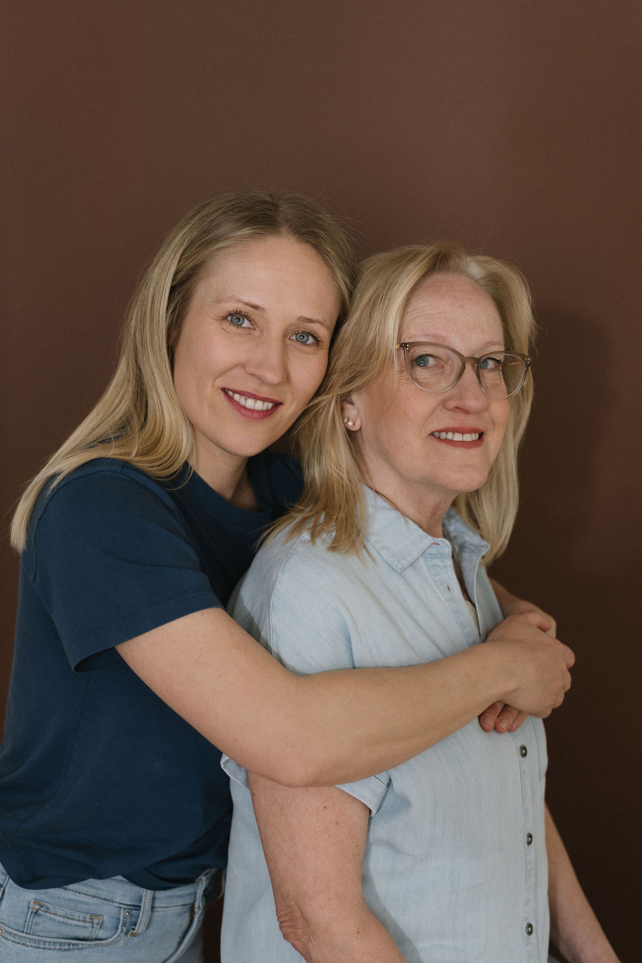 Two women, one older with glasses and the other younger with blonde hair, are smiling and hugging each other against a solid brown background.