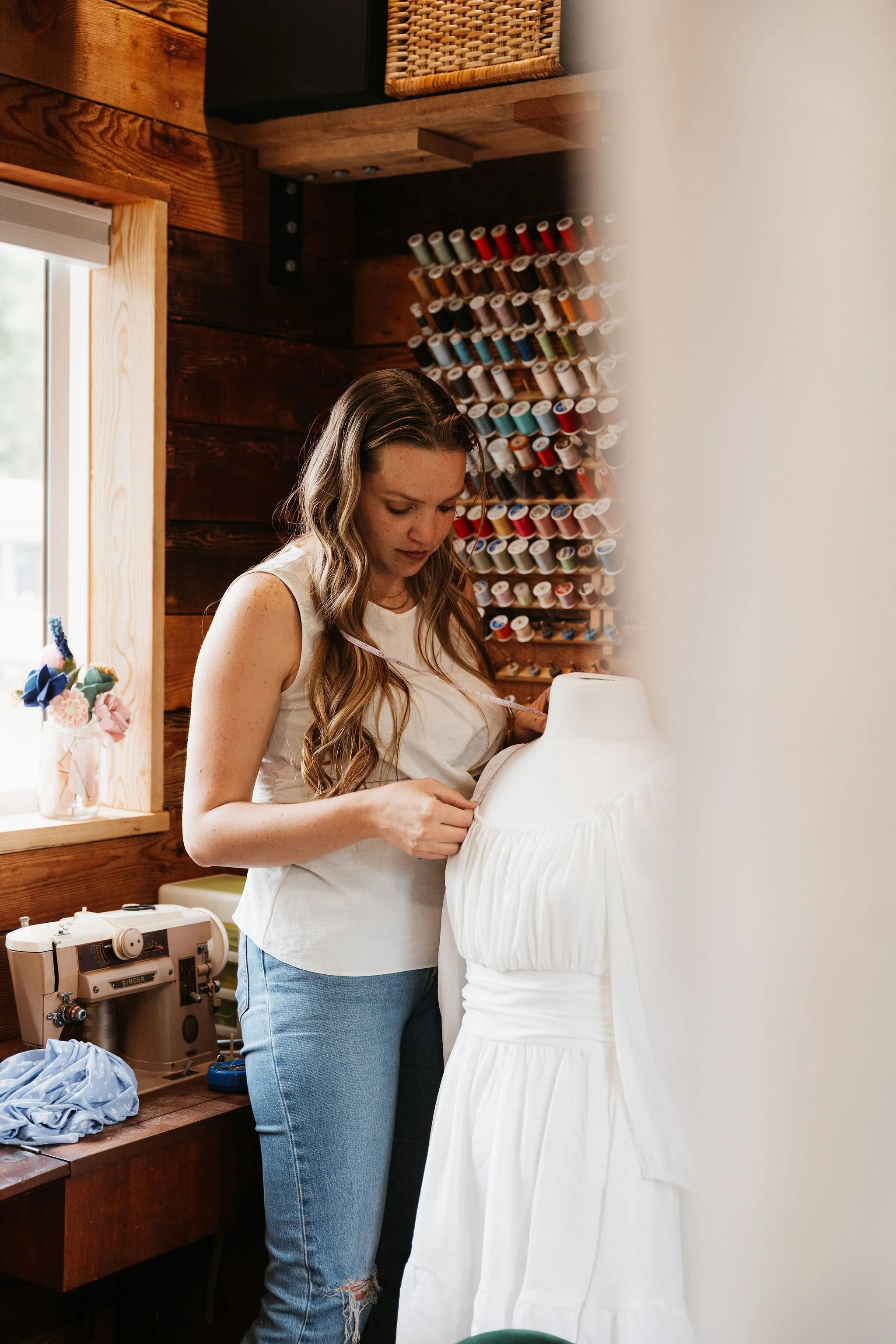 A woman in a white tank top and jeans measuring a white dress on a mannequin in a craft room with sewing supplies and thread spools on the wall.