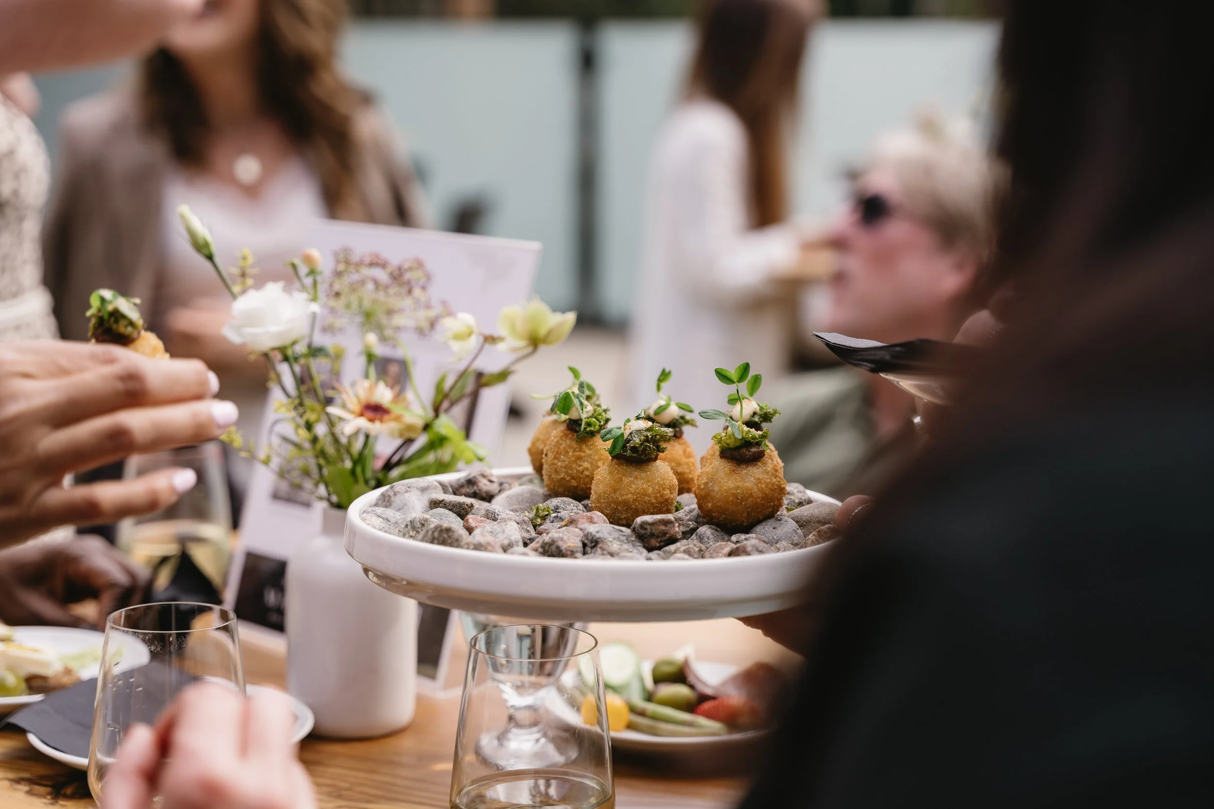 People gathered around a table enjoying appetizers and drinks, with a focus on a platter of fried food topped with herbs in the center.