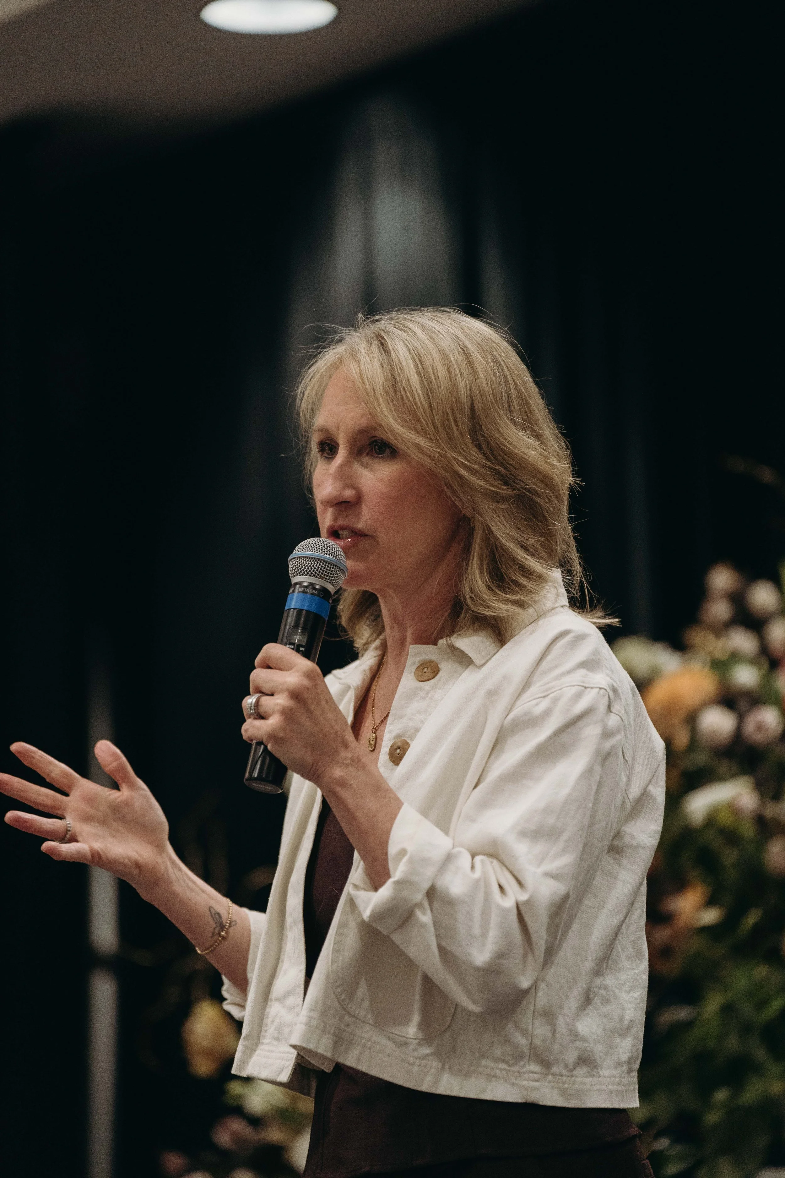 A woman with blonde hair is speaking into a microphone, gesturing with her right hand, in a setting with dark curtains and blurred flowers in the background.